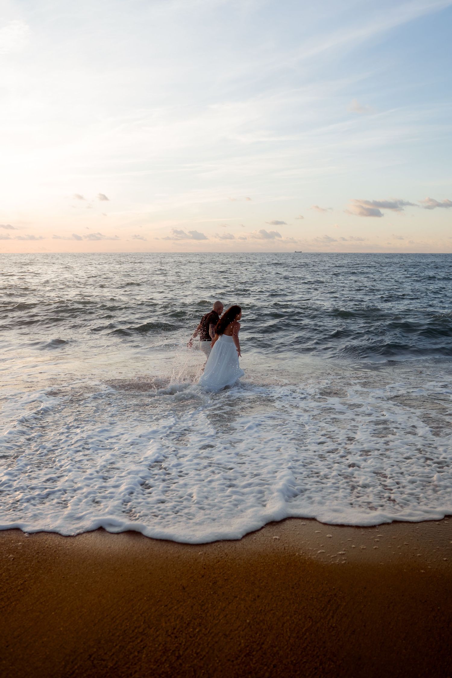Woman in a white dress wading into ocean waves alone on a beach during a colorful sunset.