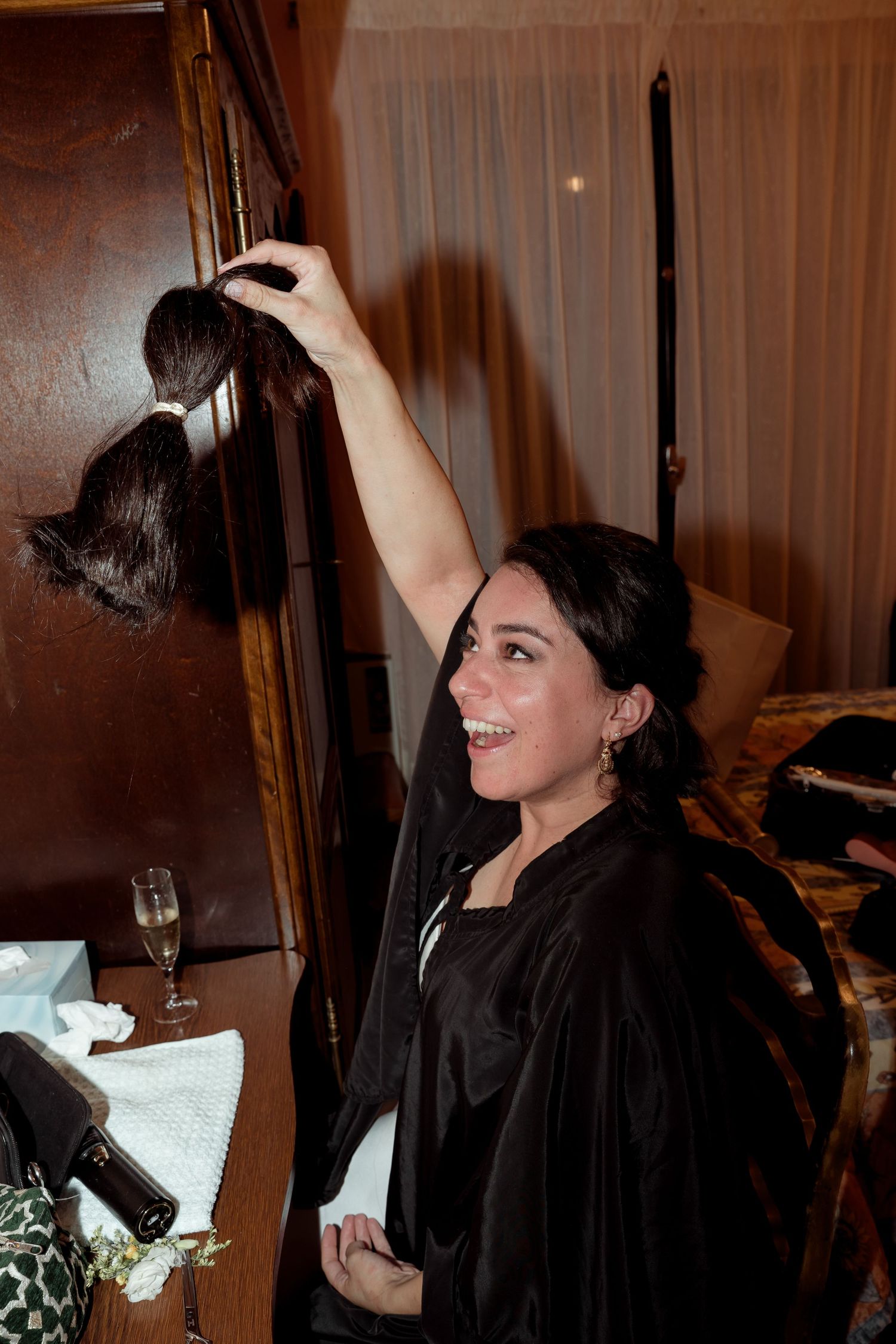 Smiling bride in black robe holds up hair extension while getting ready for wedding.