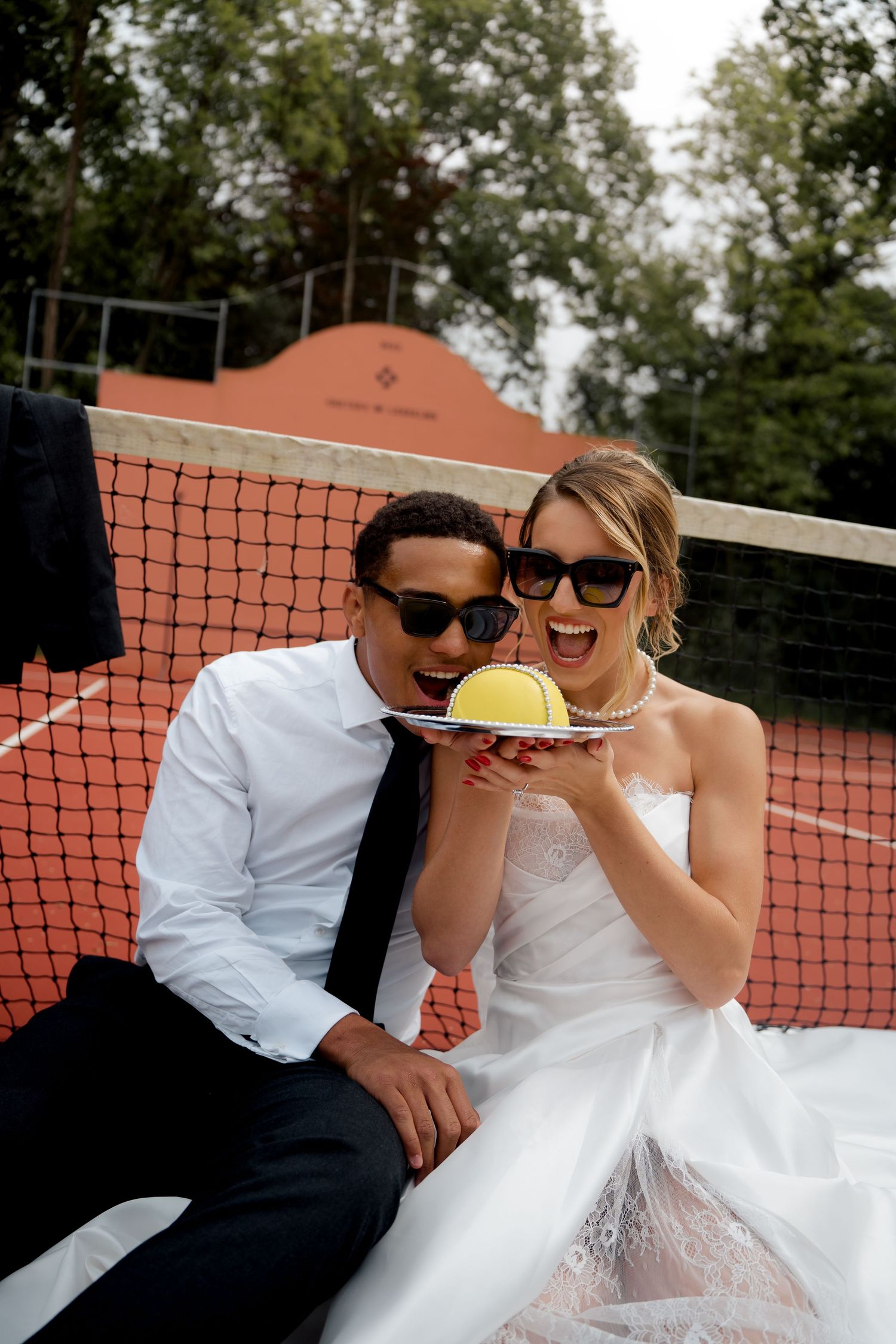 Bride and groom in sunglasses laugh while presenting a tennis ball on a plate courtside.