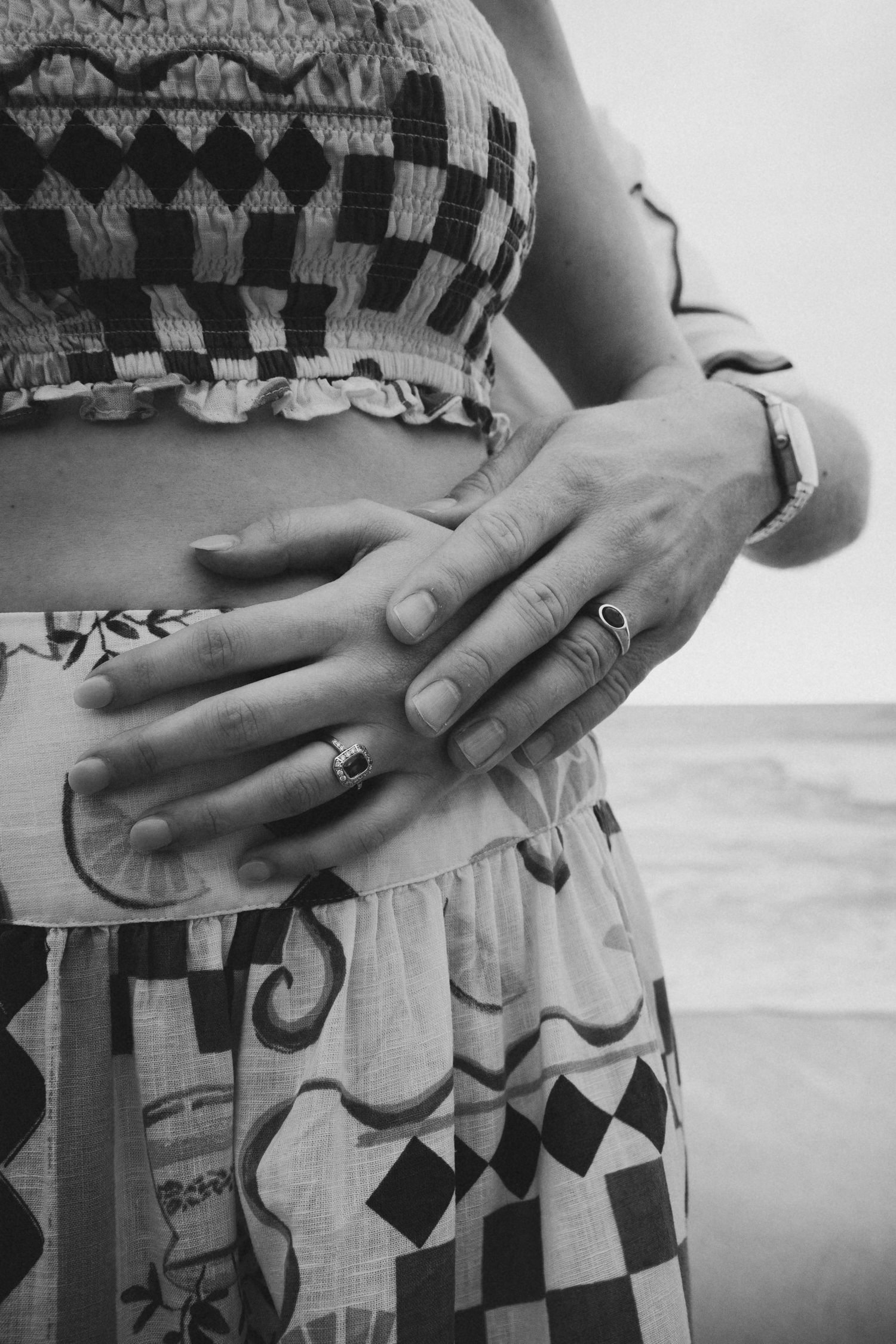 Close-up black and white photo of couple's hands with rings on woman's waist at beach.