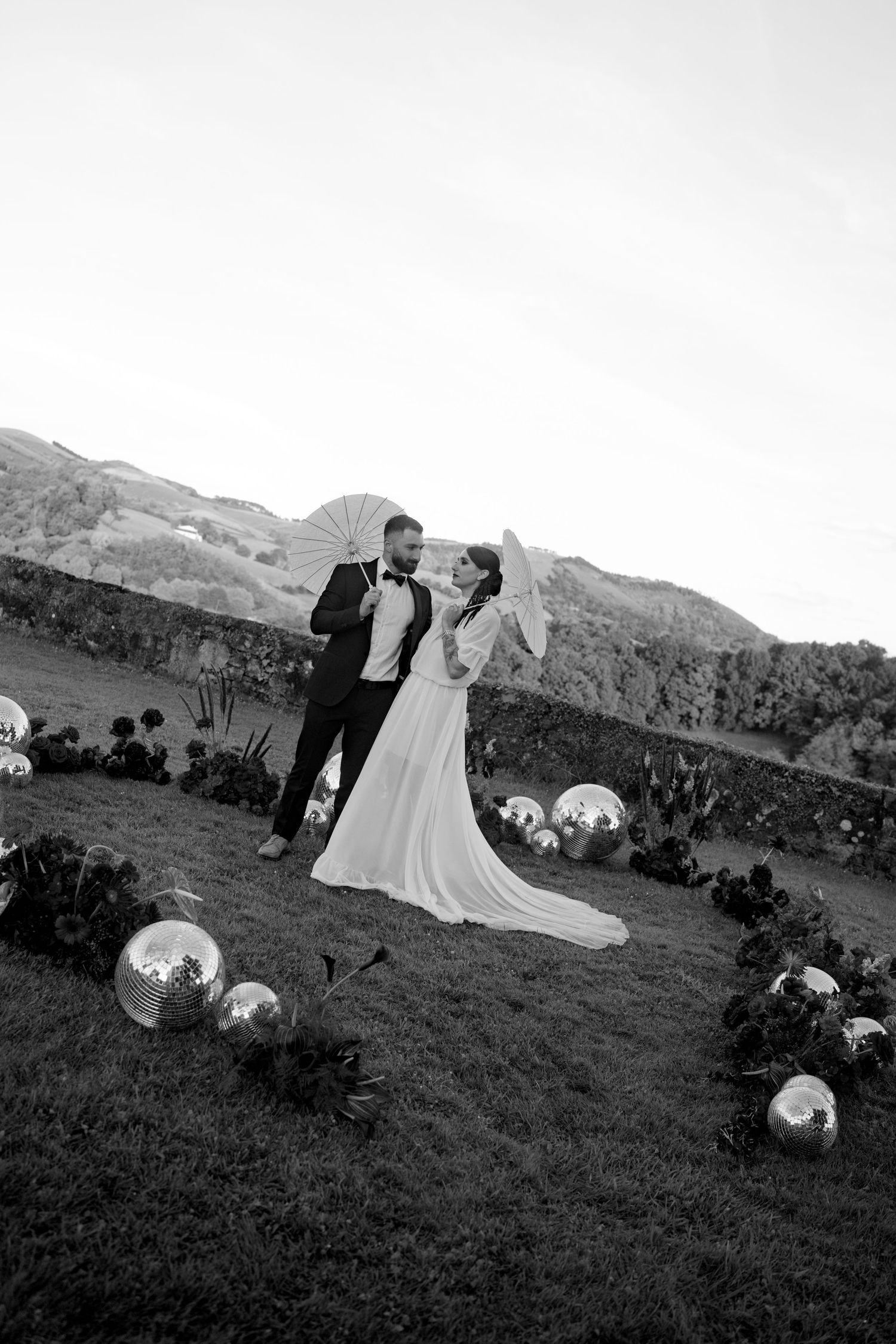 Black and white photo of bride and groom posing with white parasols along a disco ball lined aisle.