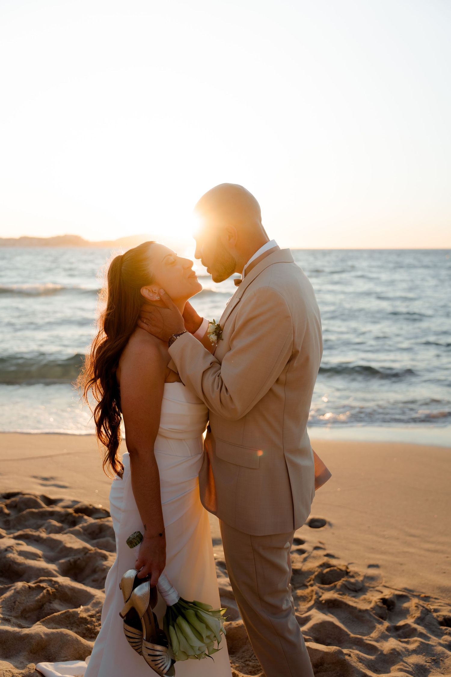 Couple sharing intimate moment on beach at golden sunset, bride holding white flower bouquet.