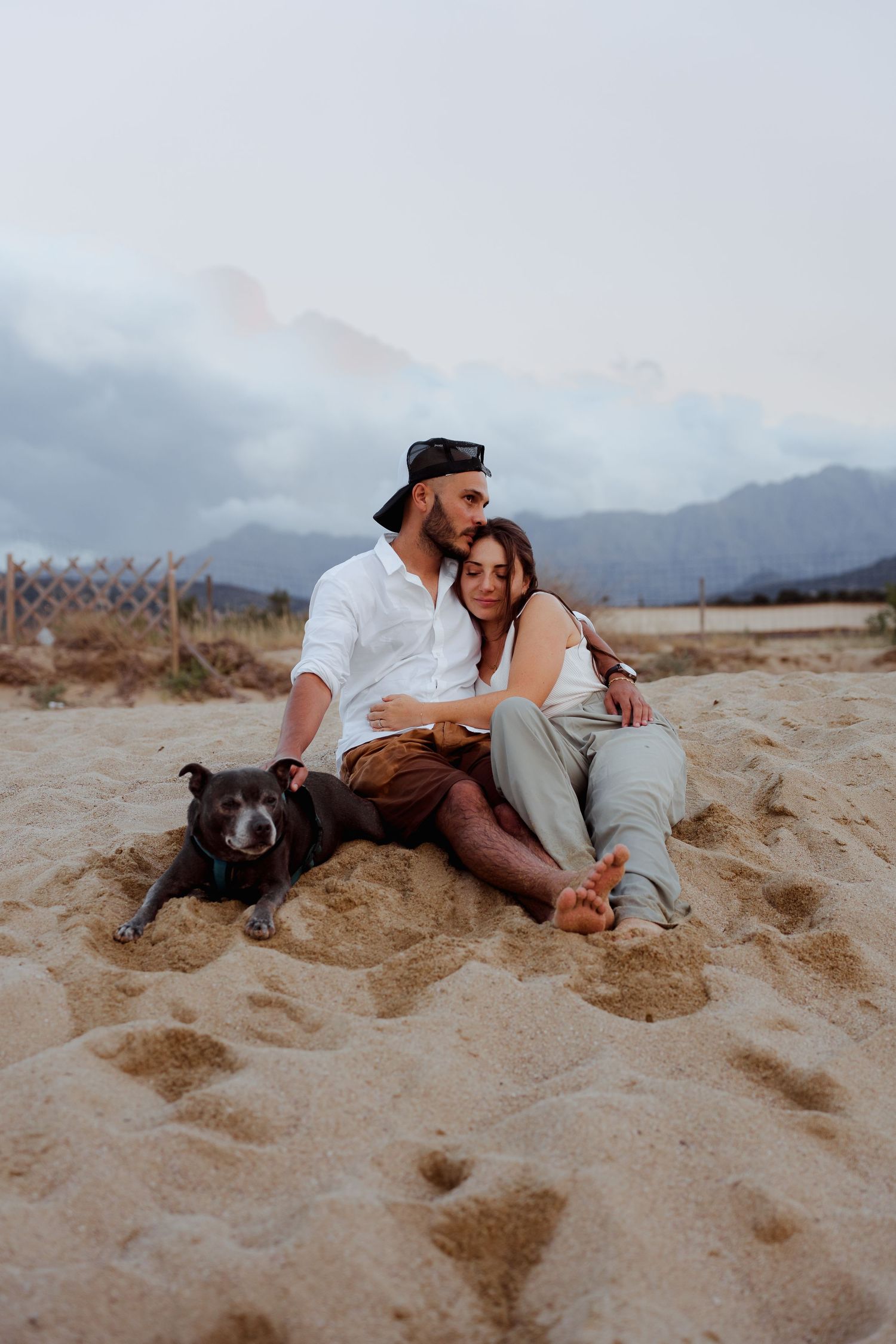 Couple sitting together on a sandy beach with their black dog, mountains and cloudy sky in background.