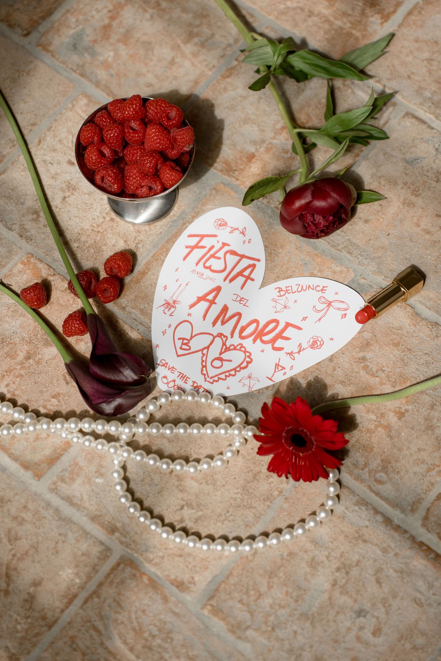 Heart-shaped Fiesta Amore card surrounded by roses, raspberries, pearl necklace, and lipstick on terracotta tiles.