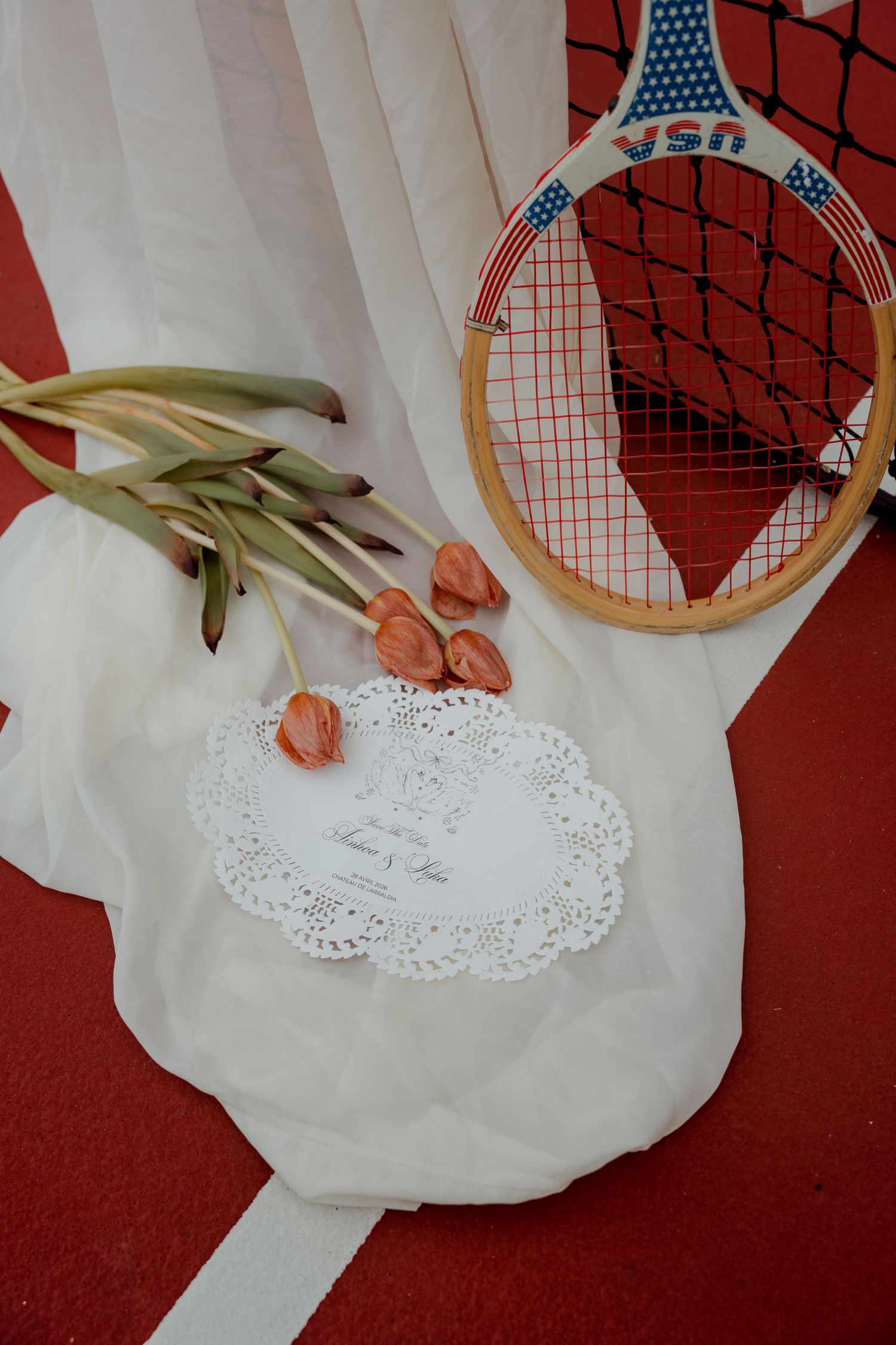 Vintage USN tennis racket beside pink tulips and lace wedding save the date card on white fabric.
