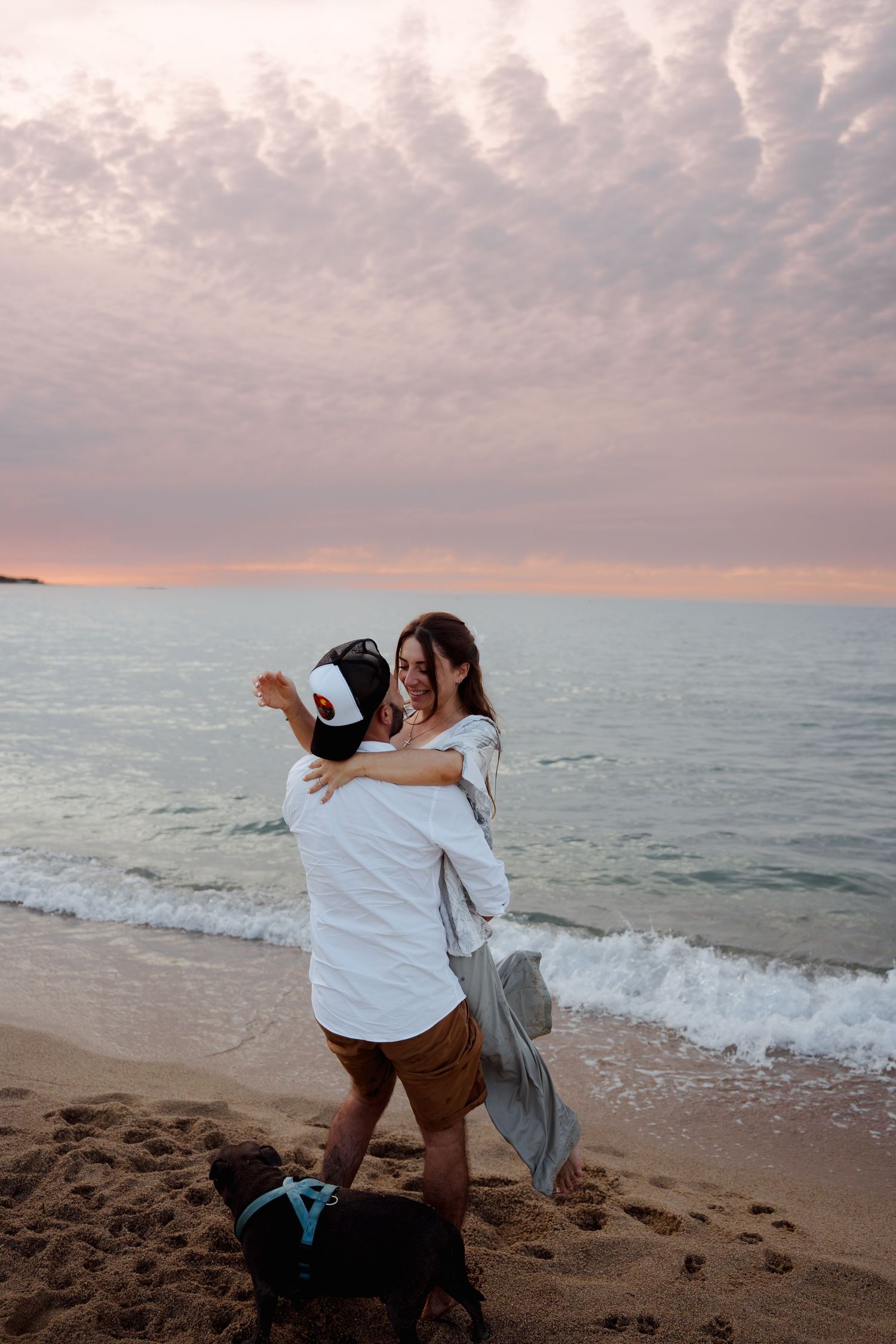 Couple embracing on a sandy beach at sunset with a dog nearby and colorful cloudy sky.