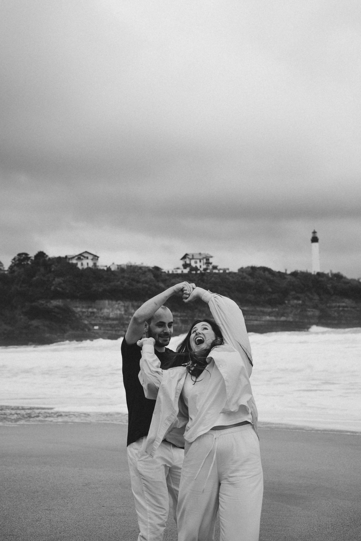 Black and white photo of a joyful couple dancing on a windy beach near a lighthouse.