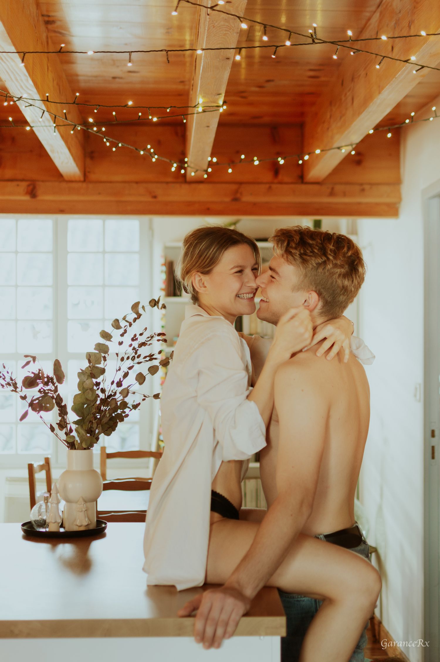 Smiling couple embracing intimately on a kitchen counter surrounded by fairy lights and dried flowers.