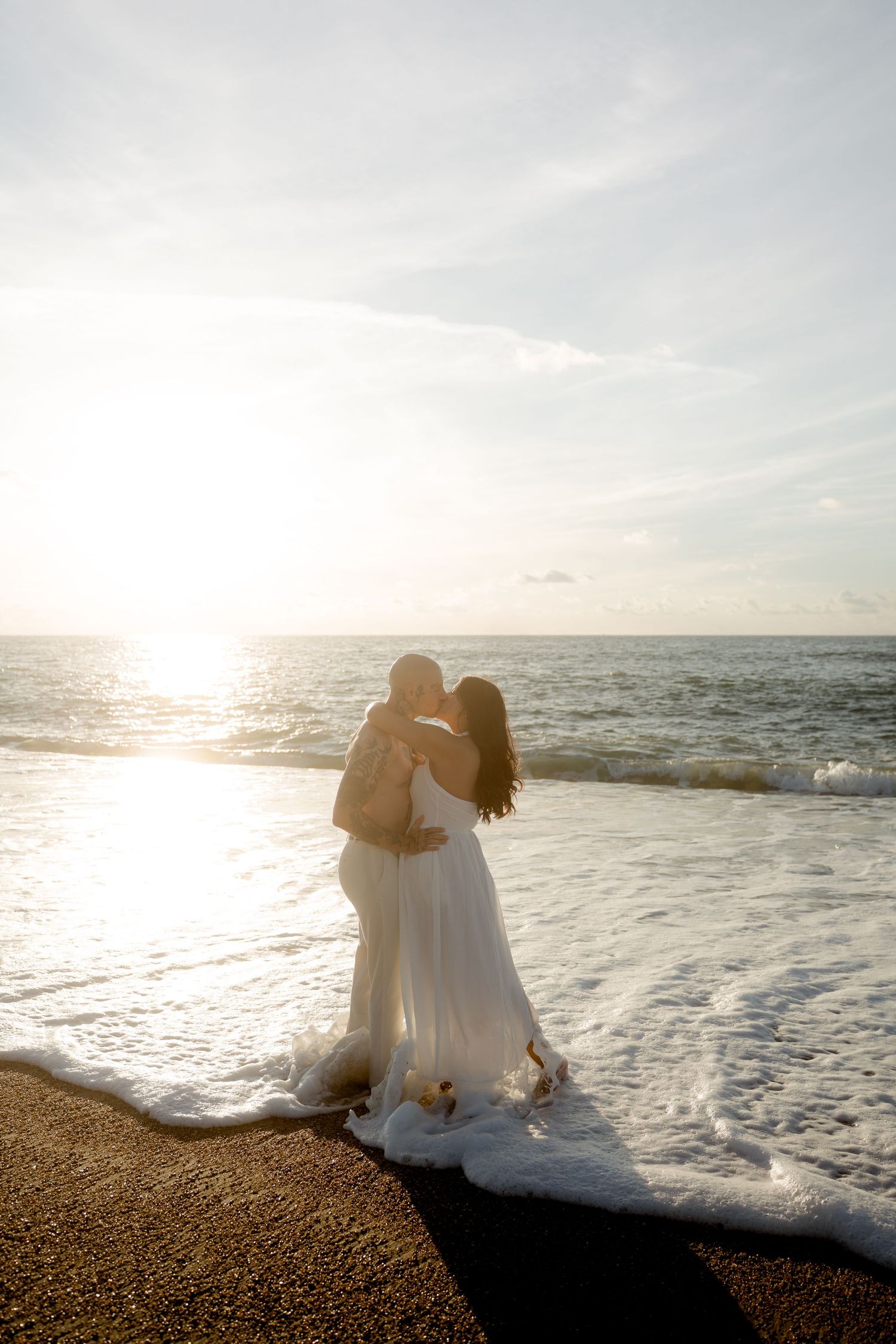 Couple kissing at water's edge during golden sunset, waves washing around woman's white flowing dress.