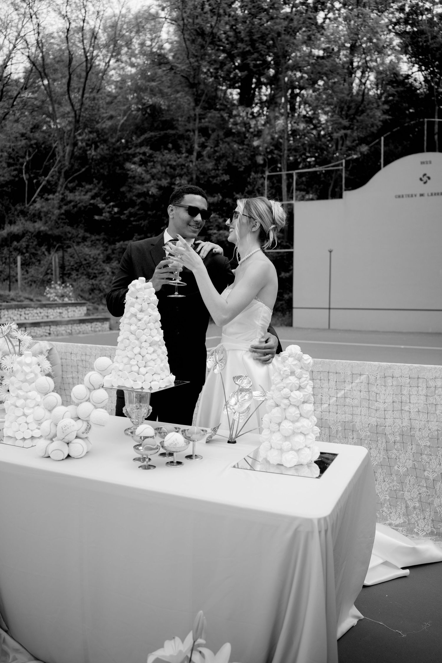 Black and white photo of bride and groom toasting champagne at macaron tower dessert table on tennis court.