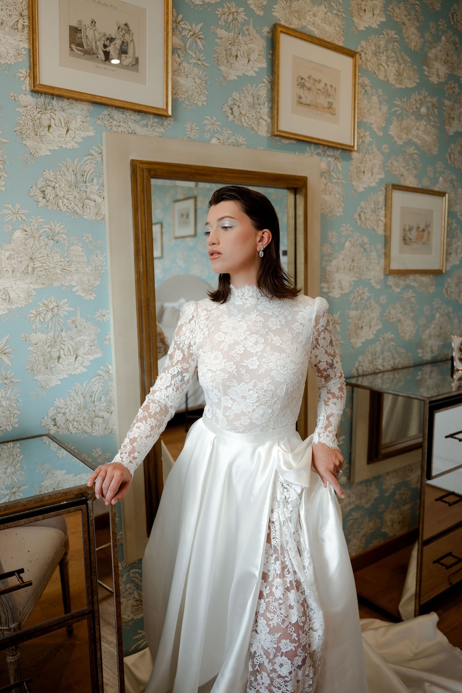 Bride in long-sleeve lace high-neck wedding dress posing elegantly in blue wallpapered dressing room.