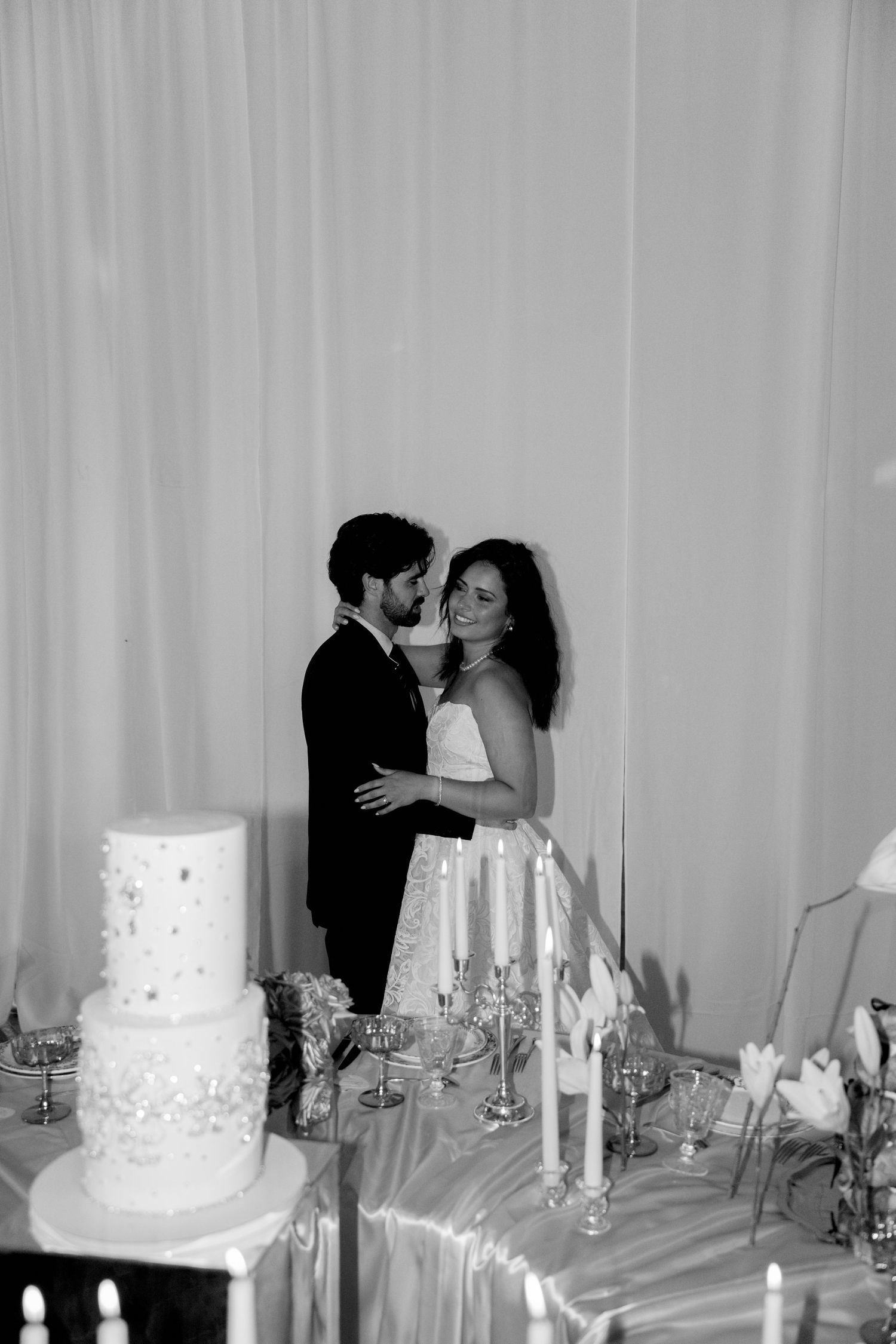 Bride and groom embracing near a tiered wedding cake and candles in a black and white photo.