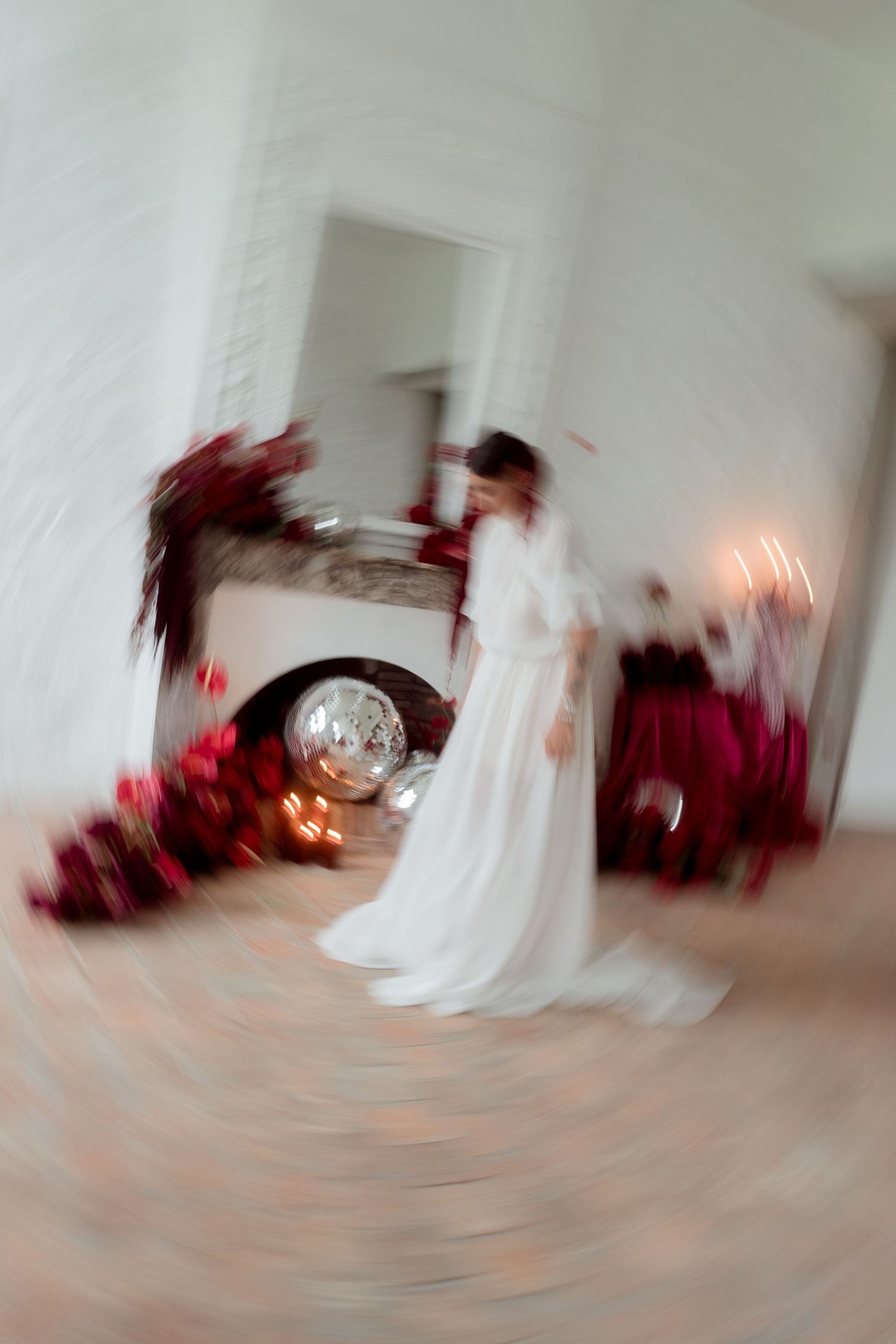 Artistic blurred motion shot of bride dancing near candlelit fireplace with red floral arrangements.