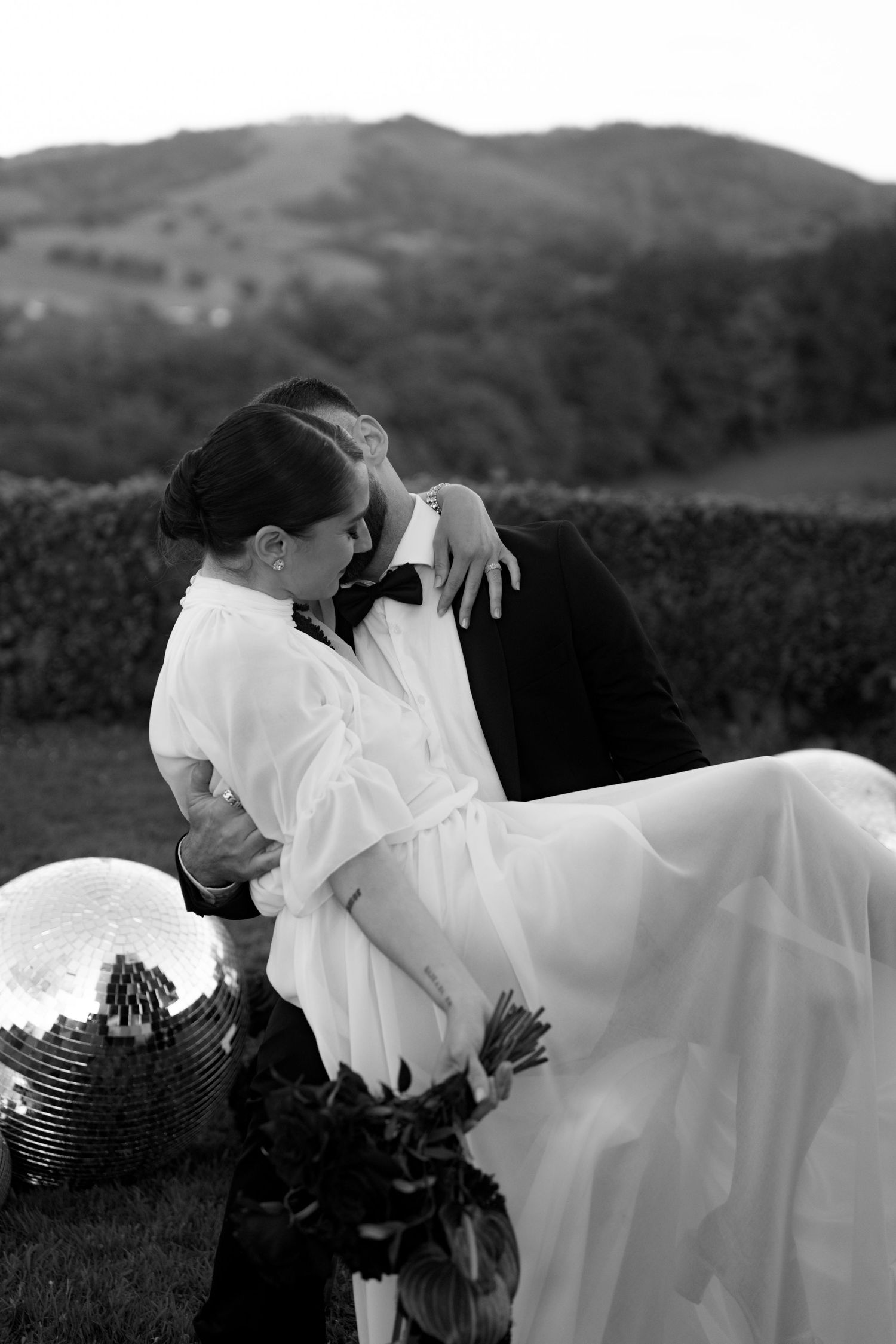 Romantic black and white wedding photo of groom holding bride near a disco ball with mountain backdrop.