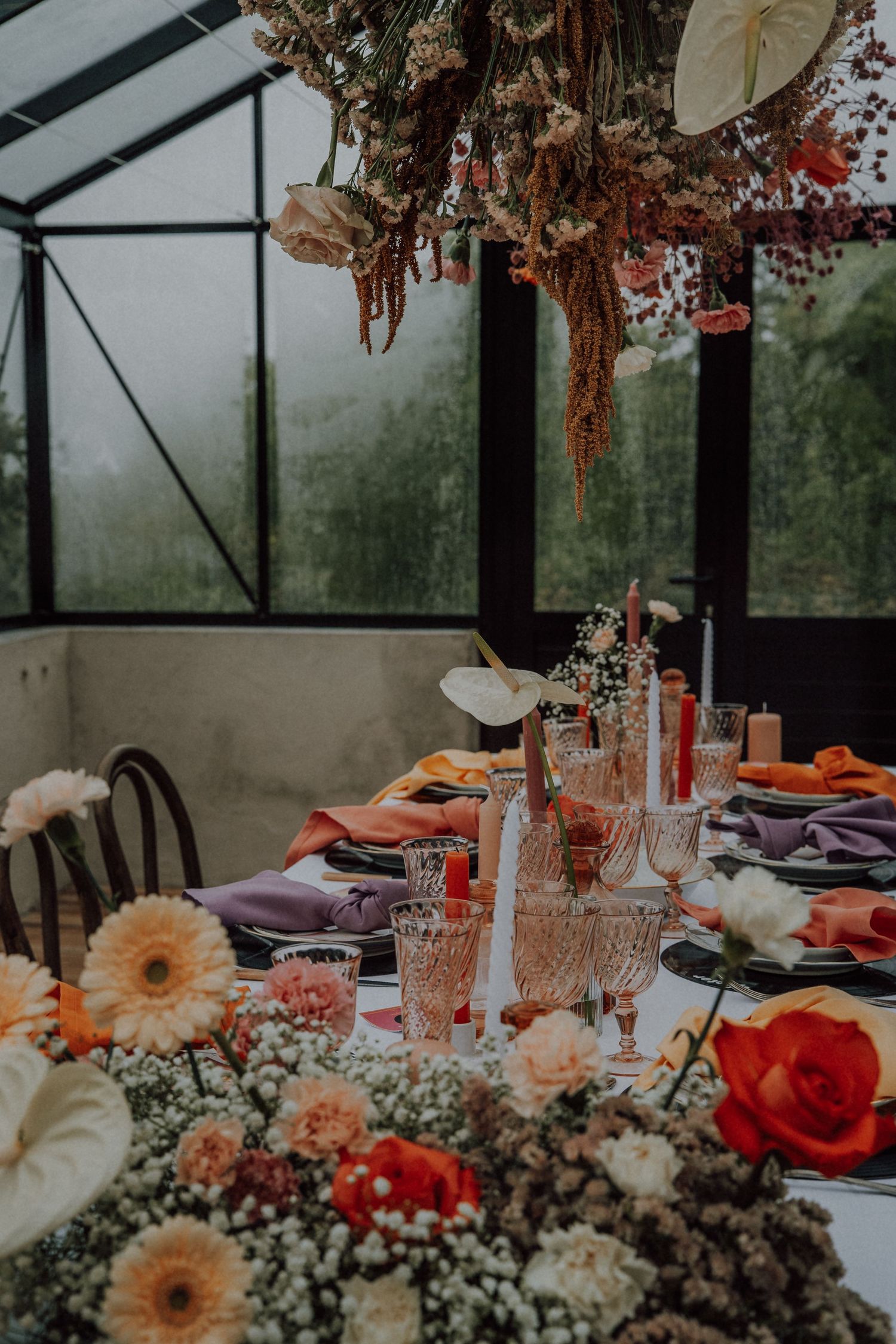 Elegant greenhouse wedding table setting with colorful flowers, pink glassware, and orange and purple napkins.