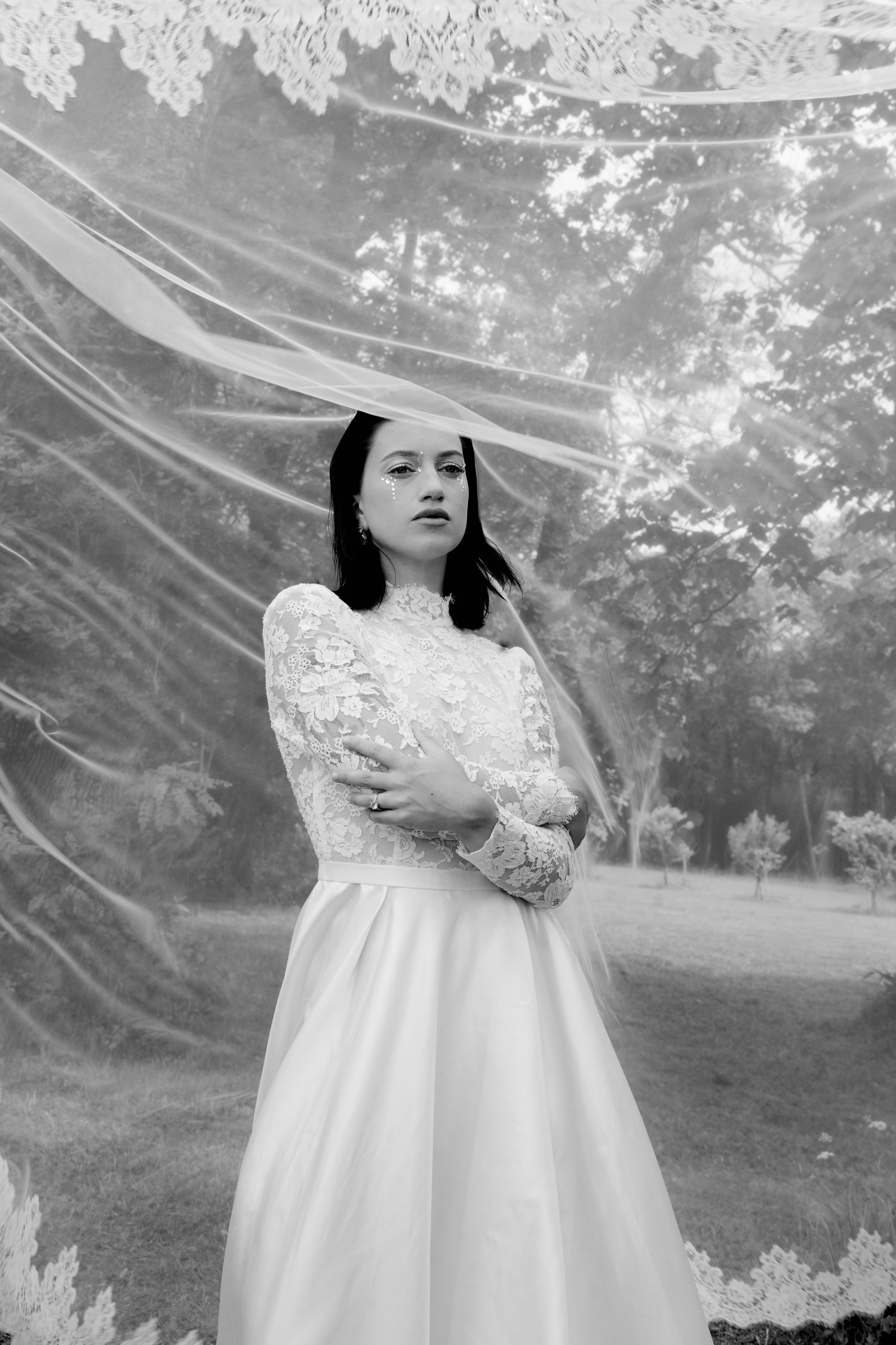 Black and white portrait of a bride in lace gown beneath a flowing veil outdoors.