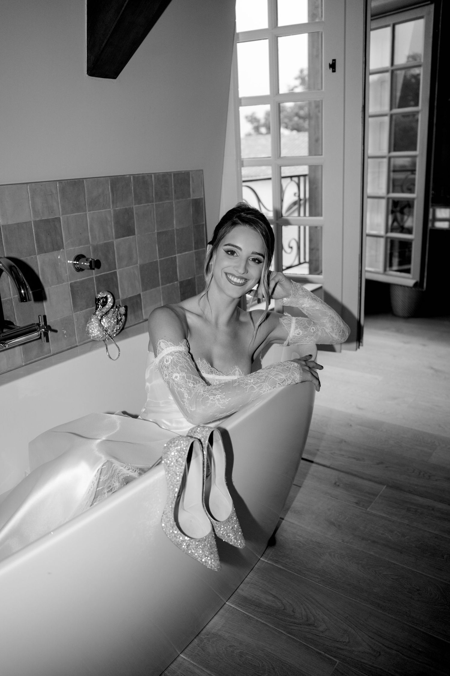 Smiling bride in lace wedding dress posing in bathtub with heels, black and white photo.