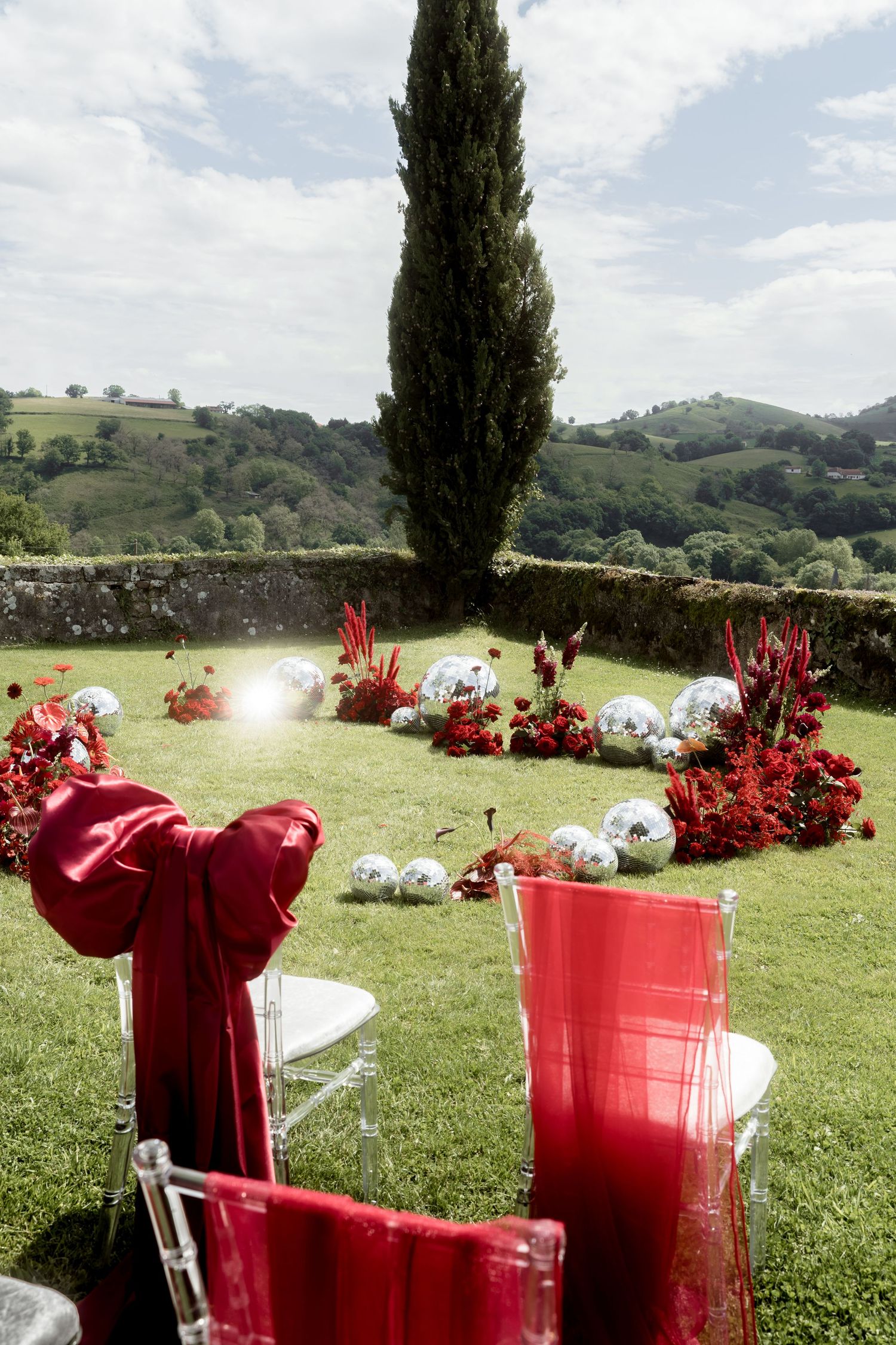 Red-draped wedding chairs with disco balls and floral arrangements on lawn beside tall cypress tree.