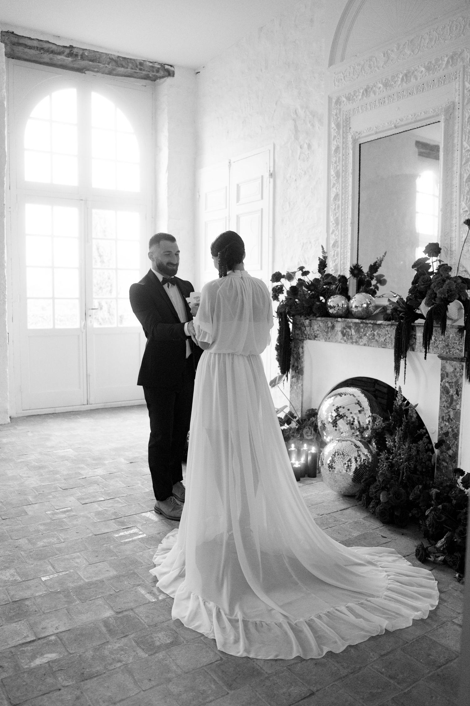 Bride and groom exchanging vows near fireplace in bright white room with dramatic floral arrangements.