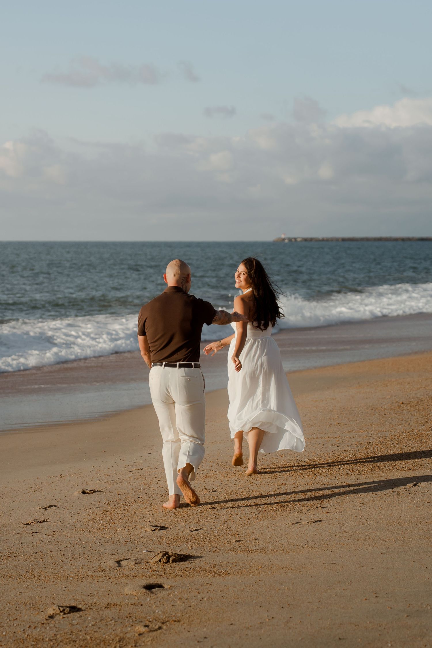 Couple running barefoot on sandy beach at sunset, woman in white dress holding hands with man in brown shirt.