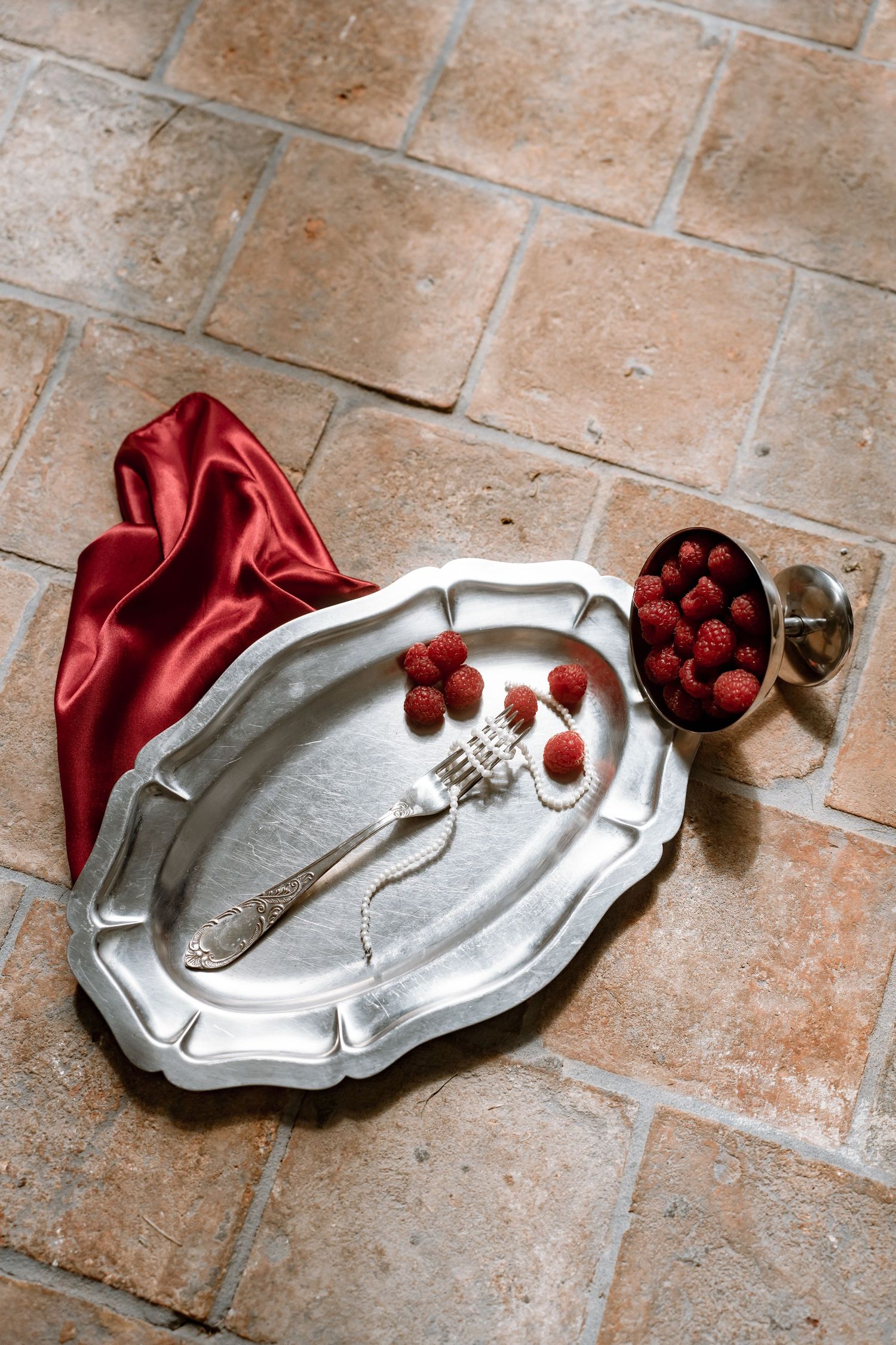Silver tray with fresh raspberries, pearl necklace, and red satin cloth arranged on terracotta tile floor.
