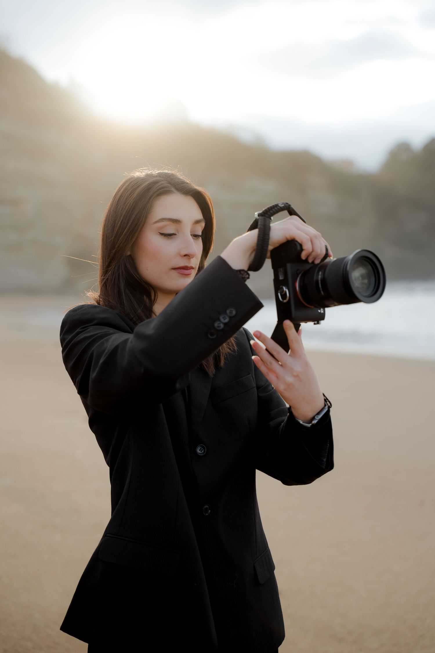 Woman in black blazer reviewing camera settings on sunlit beach at golden hour.