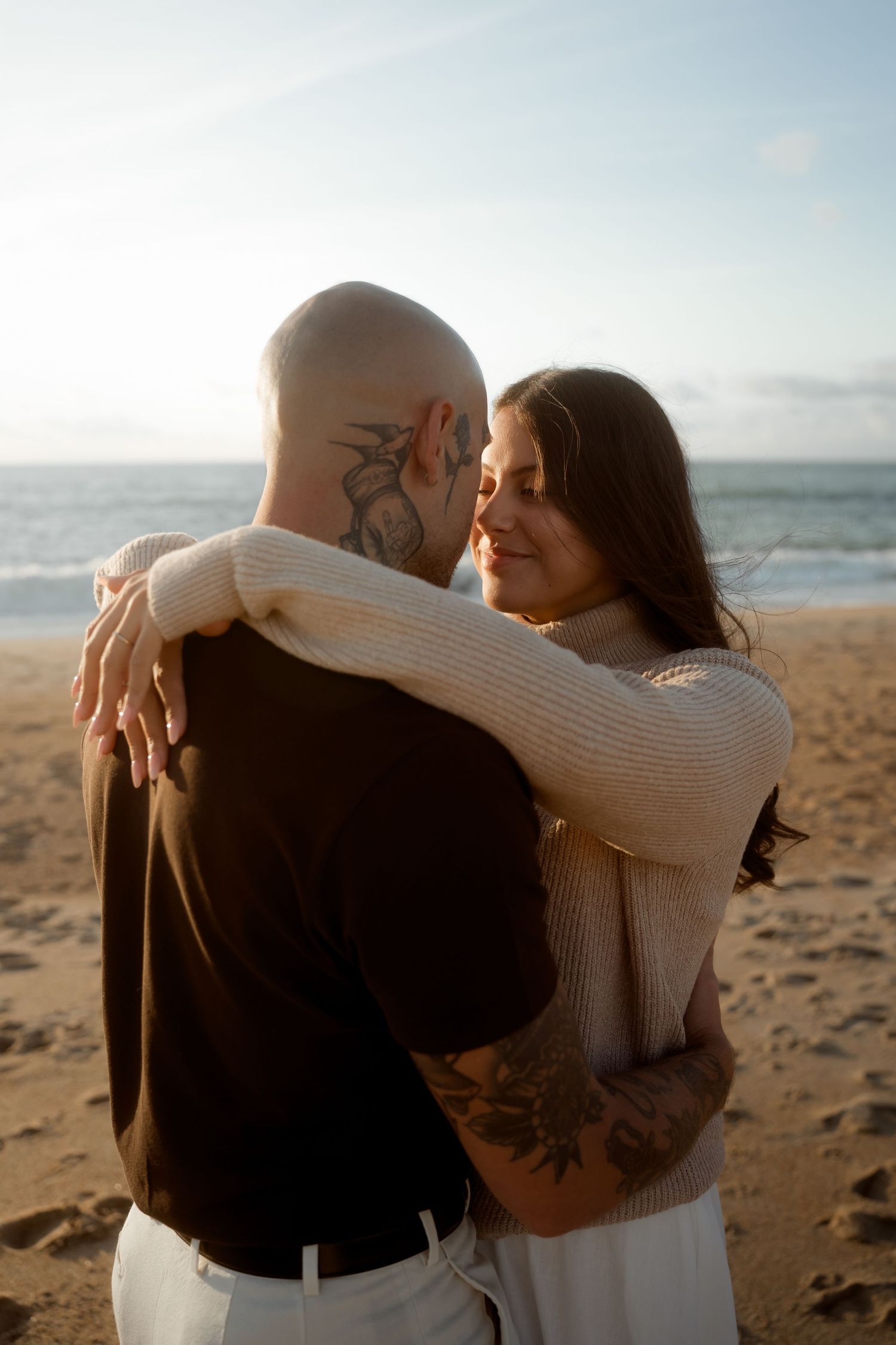 Couple embracing on a sandy beach at sunset, woman smiling with eyes closed, man with tattoos and shaved head.