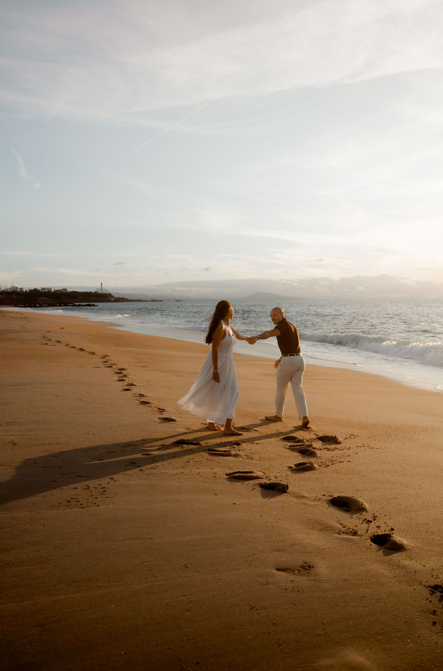 Couple dancing and holding hands on a sunlit beach at golden hour with footprints in the sand.
