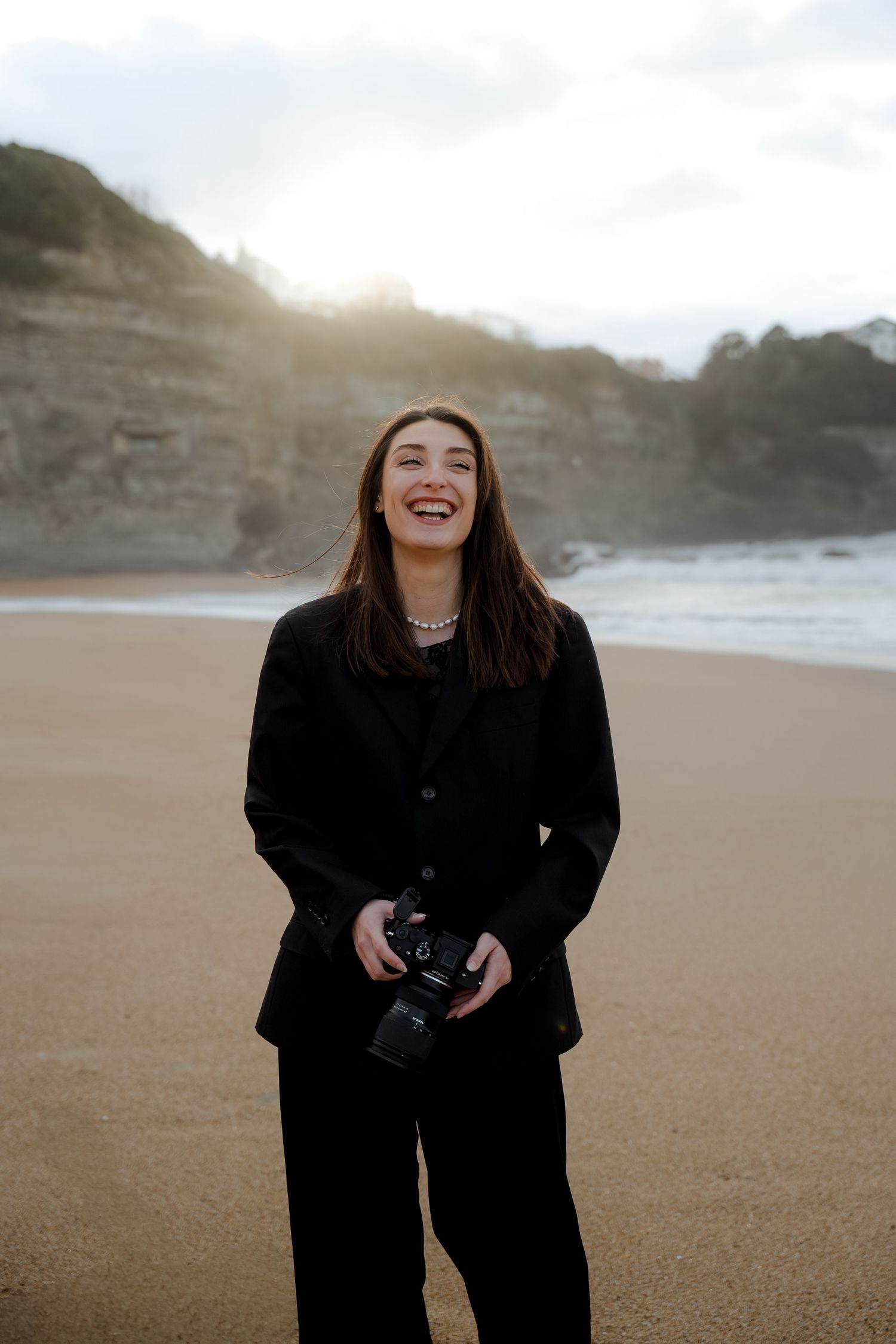 Young woman laughing on sandy beach holding DSLR camera, wearing black suit with cliffs and ocean in background.