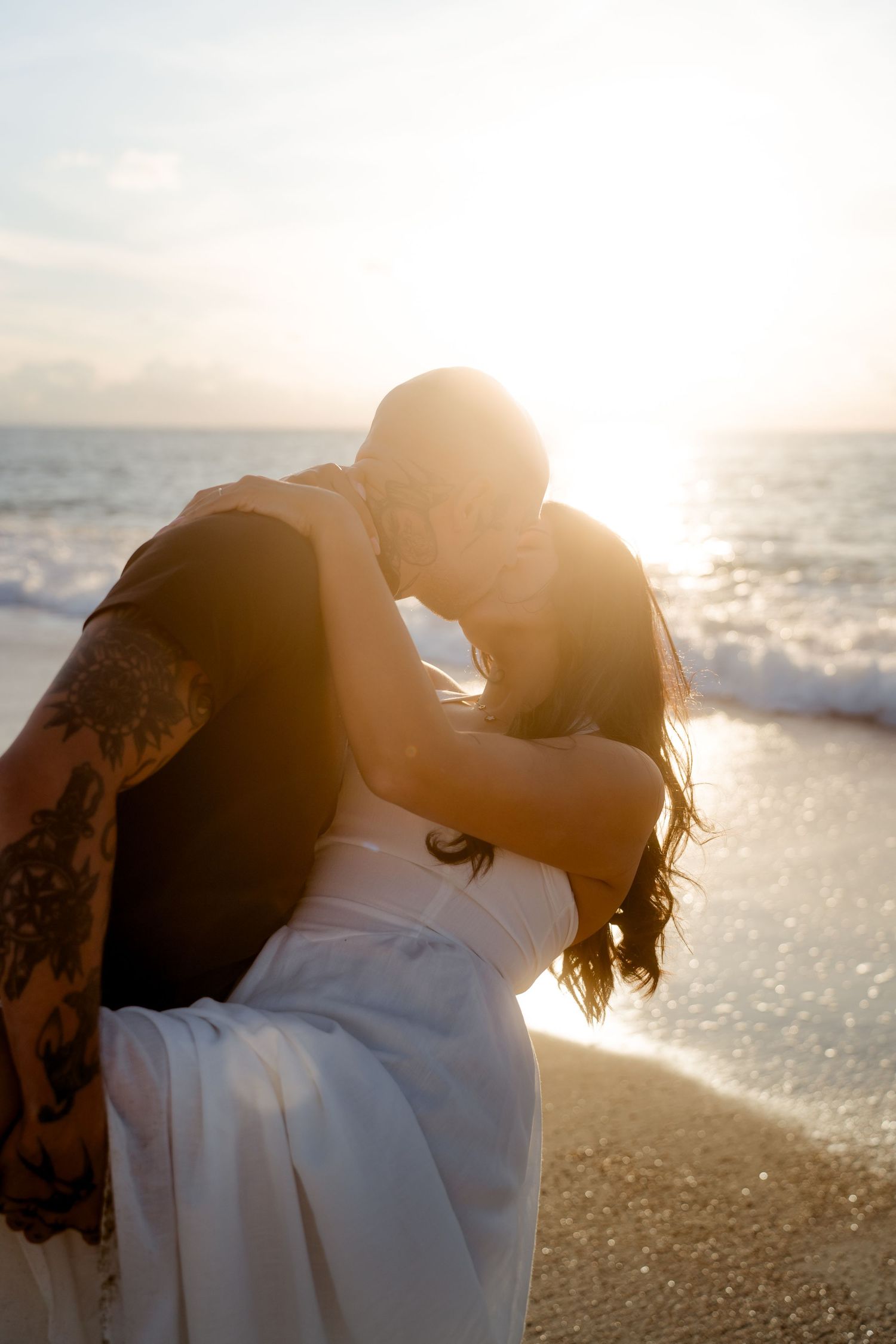 Tattooed man dipping woman in white dress for a kiss on glowing sunset beach.