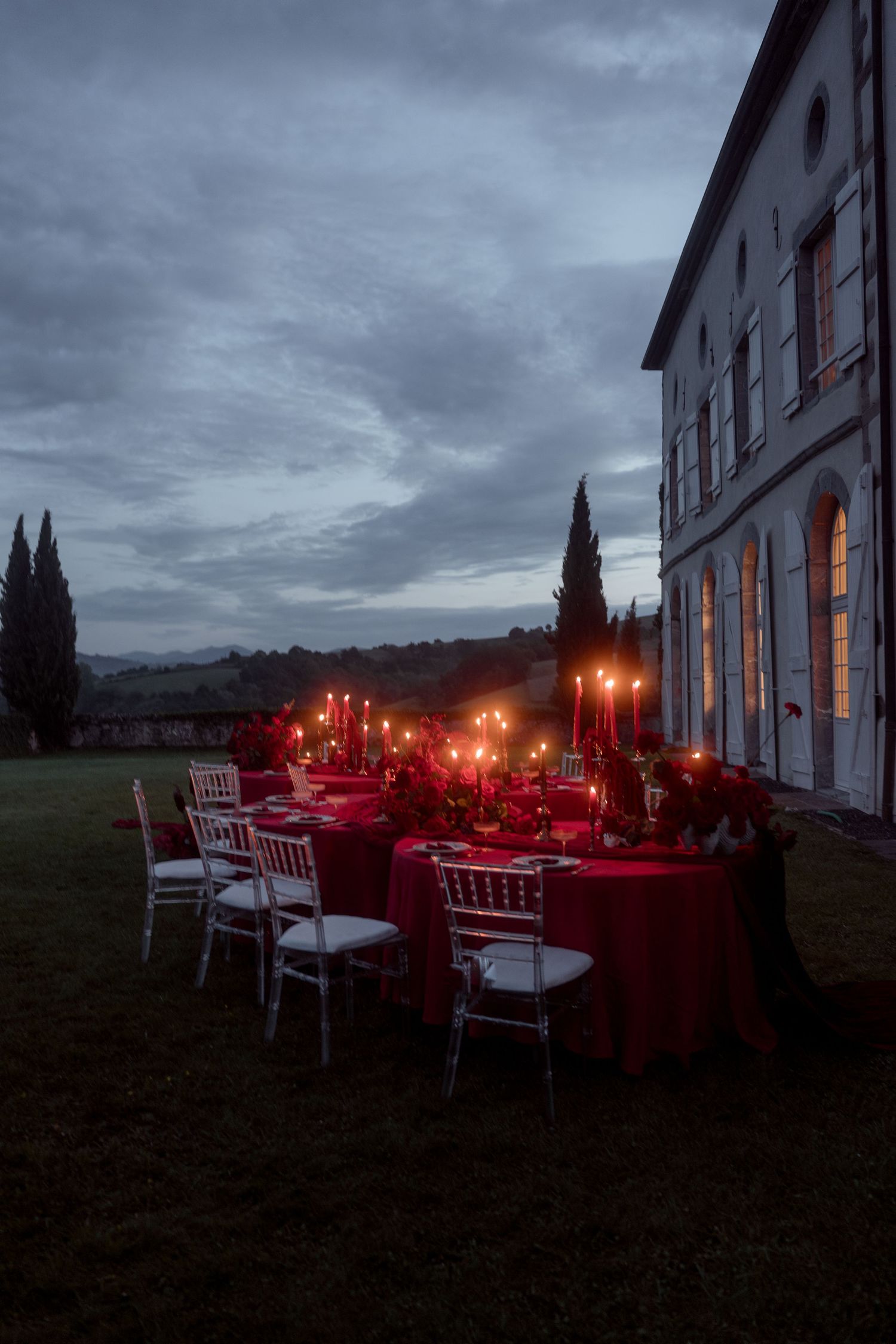 Romantic outdoor wedding reception with red-draped tables and candlelight beside illuminated Italian villa at twilight.