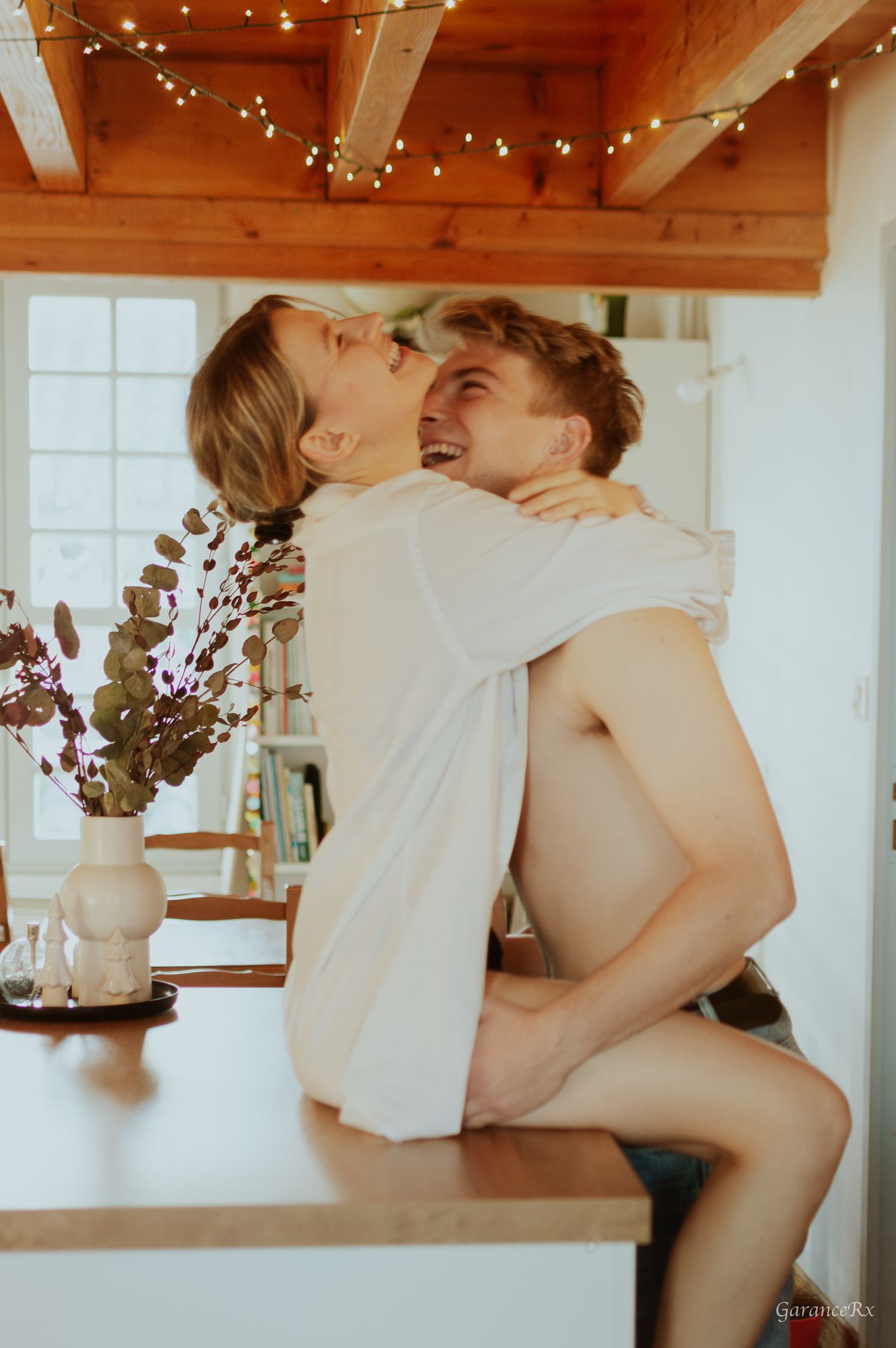 Joyful couple laughing and hugging on a kitchen counter beneath glowing string lights indoors.