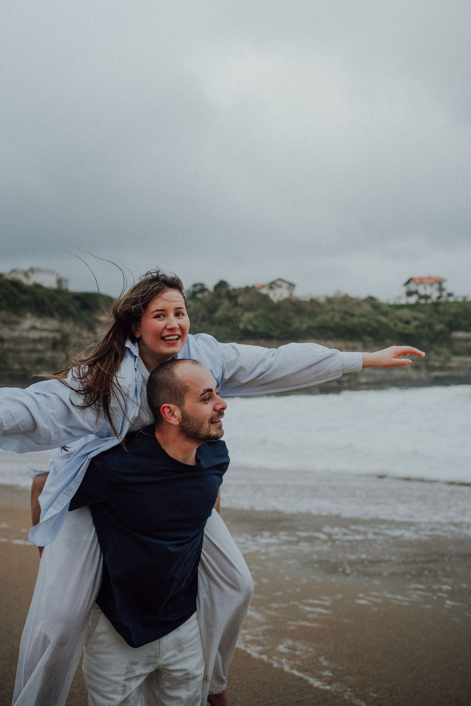Laughing woman riding on mans shoulders at beach, arms spread wide with ocean waves and cliffs behind.