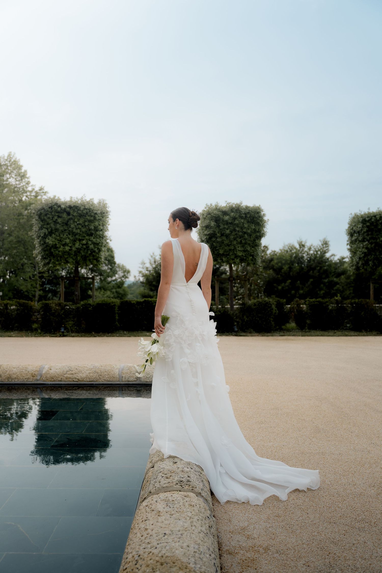 Bride in elegant backless white gown with train stands beside reflective pool in formal garden.