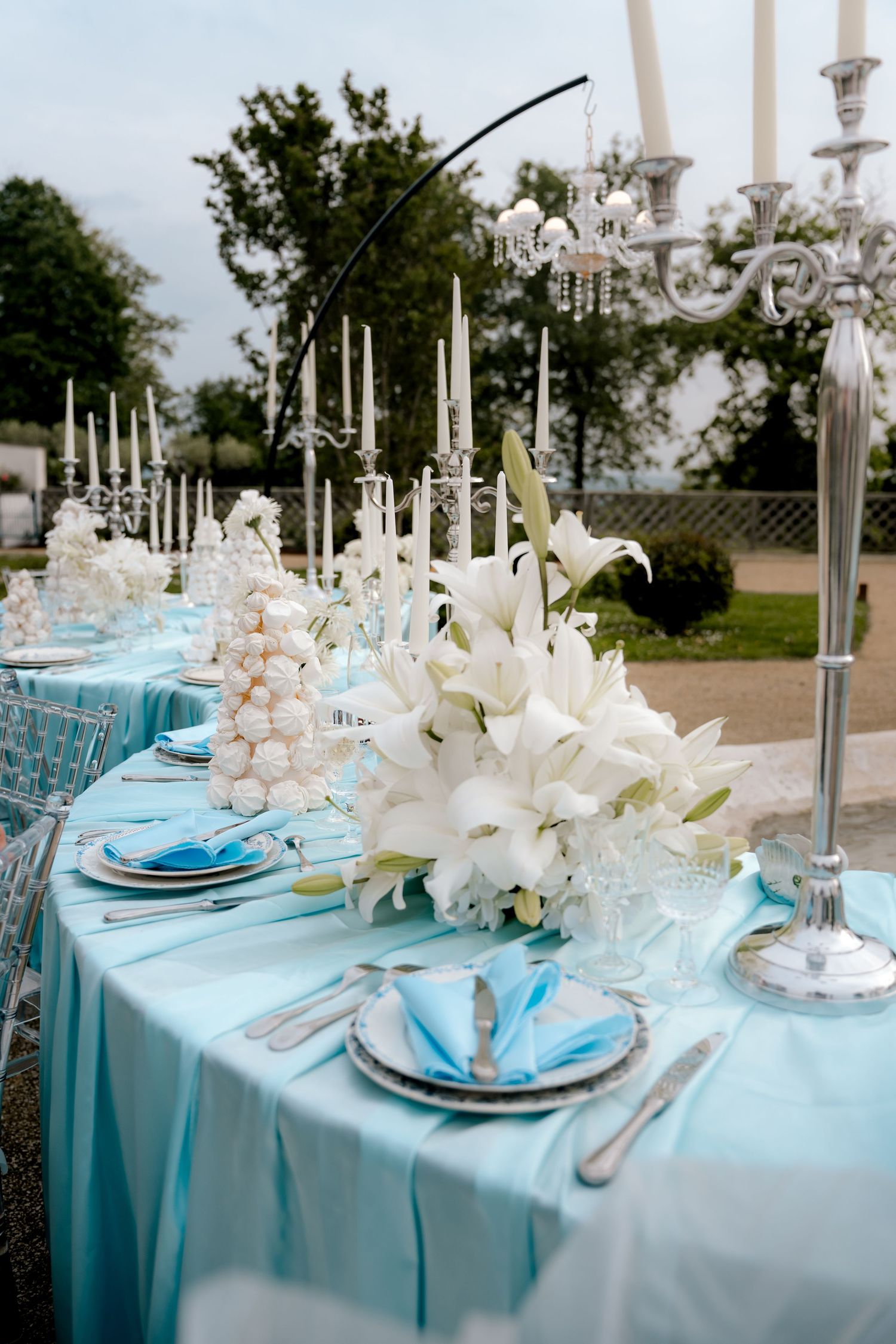 Elegant outdoor wedding reception table with blue linens, white lilies, candelabras, and crystal glassware.