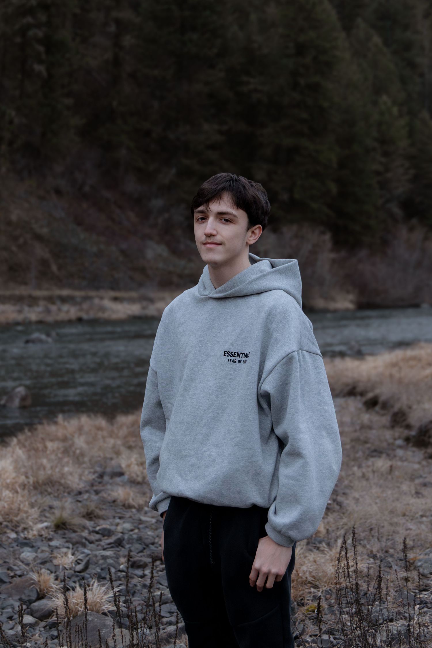 Person in a gray sweatshirt and dark pants poses in a natural outdoor setting with dry grass and trees in background.