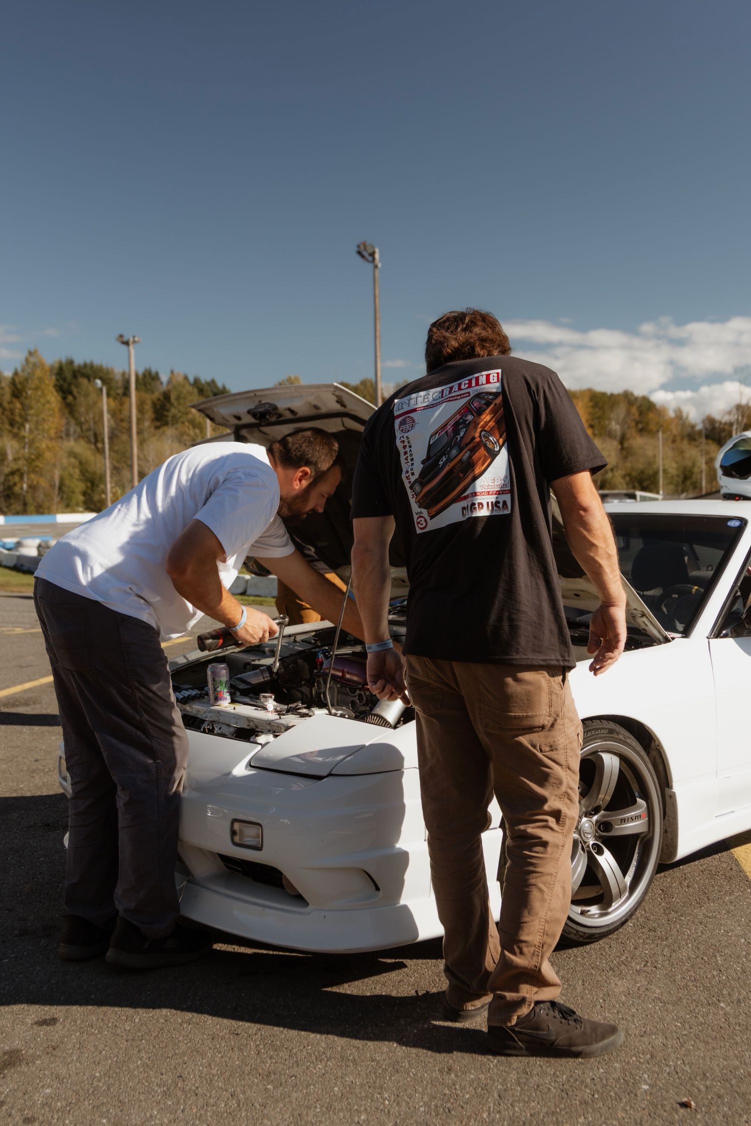 Two people examining under the hood of a white sports car in a parking lot.