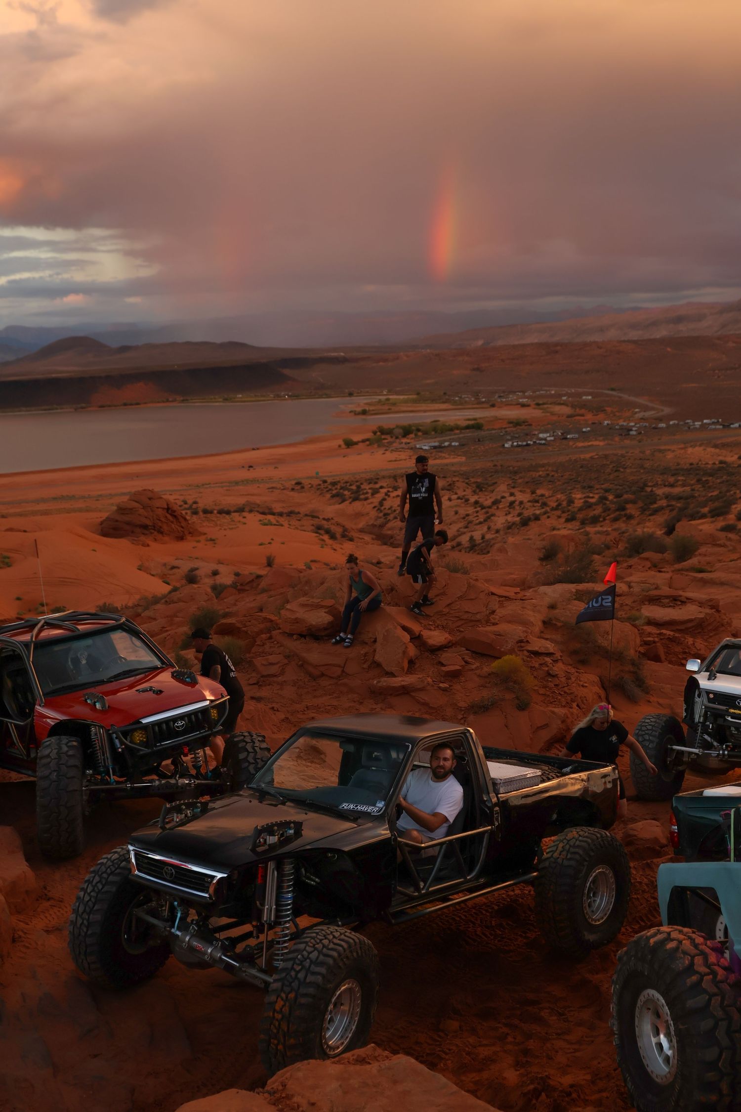 Off-road vehicles gathered at sunset on red desert terrain with rainbow in background.