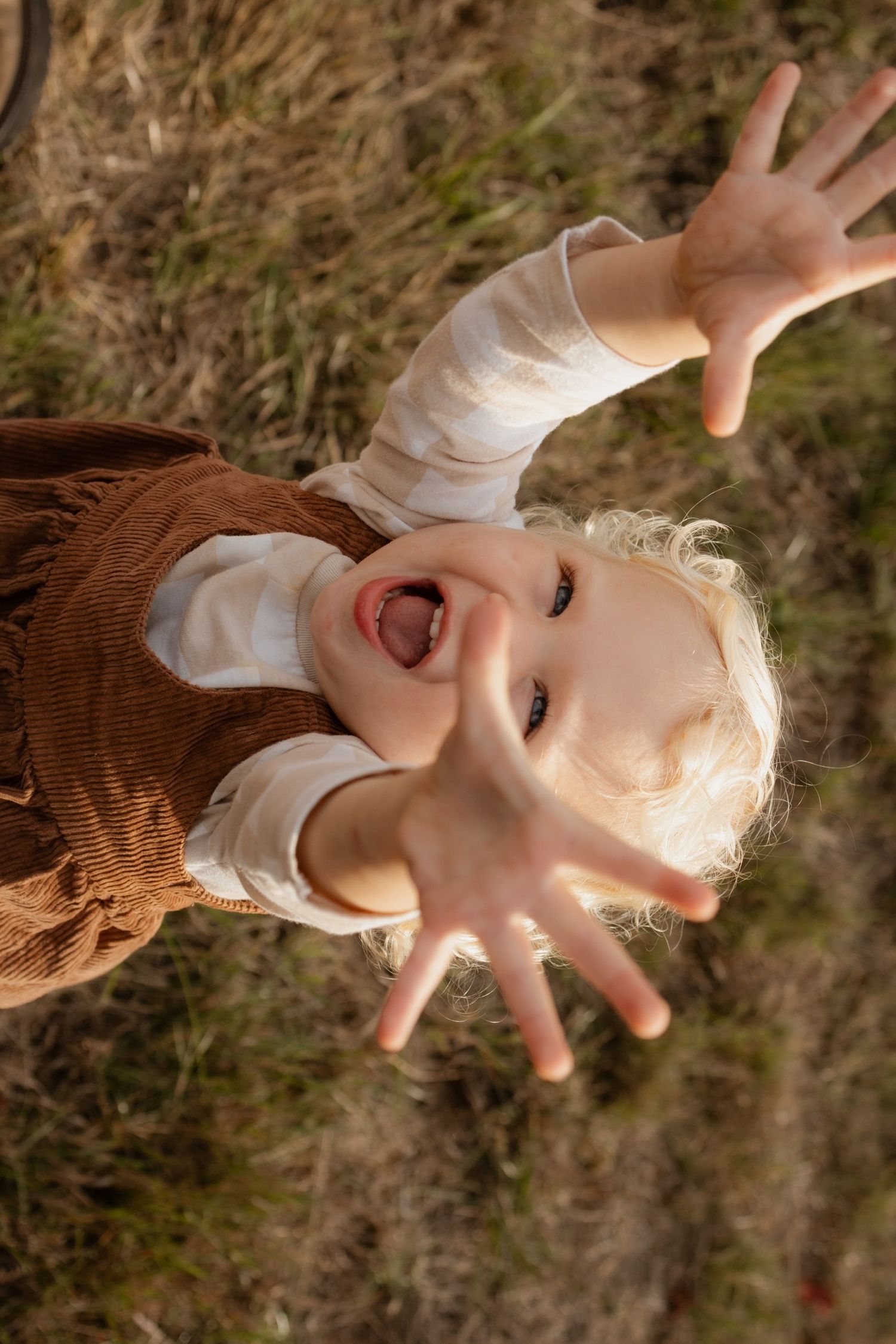 A playful child lying on grass reaching up towards camera with an excited expression.
