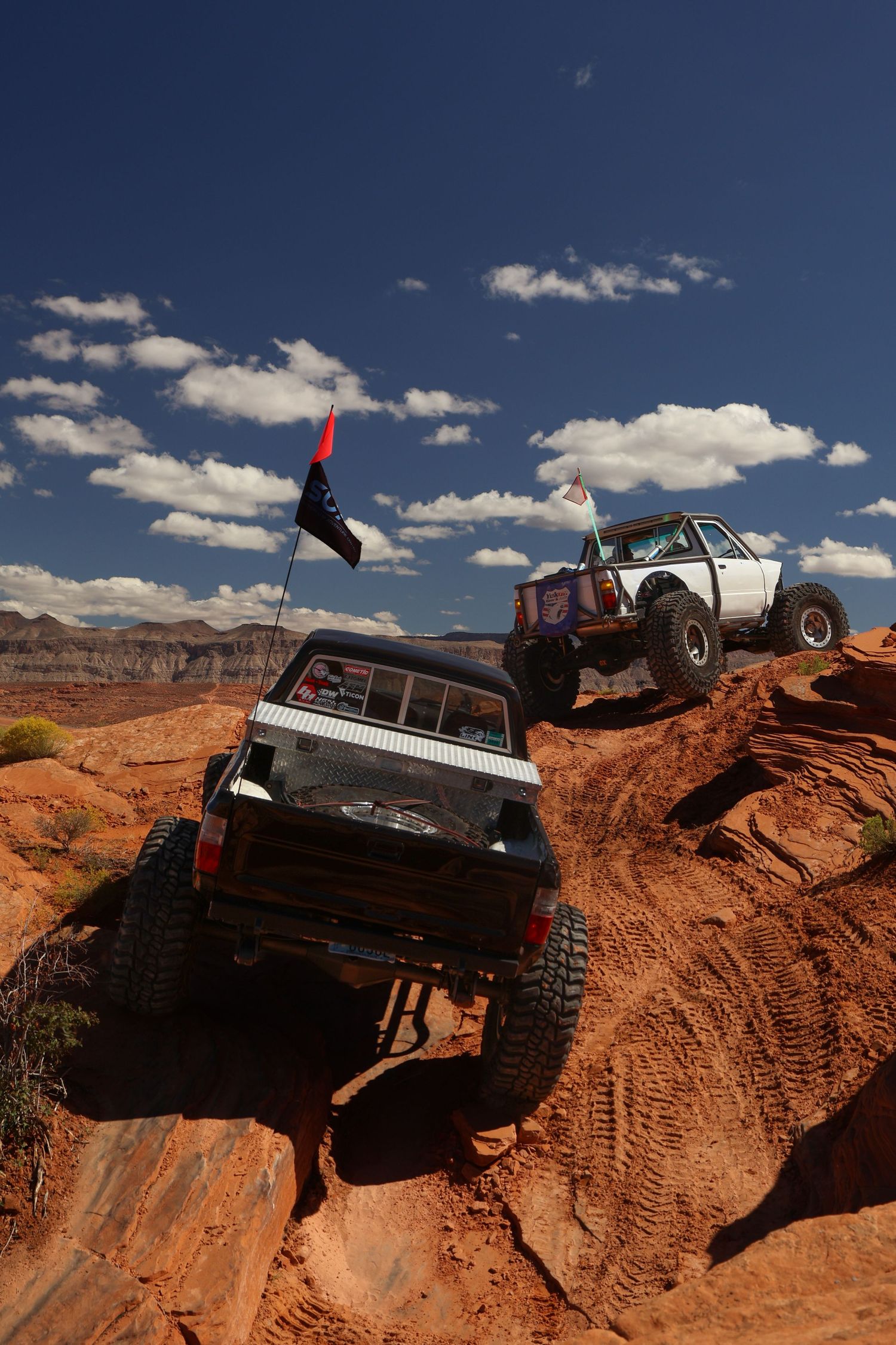 Off-road vehicles navigate steep red rock terrain against a bright blue desert sky.