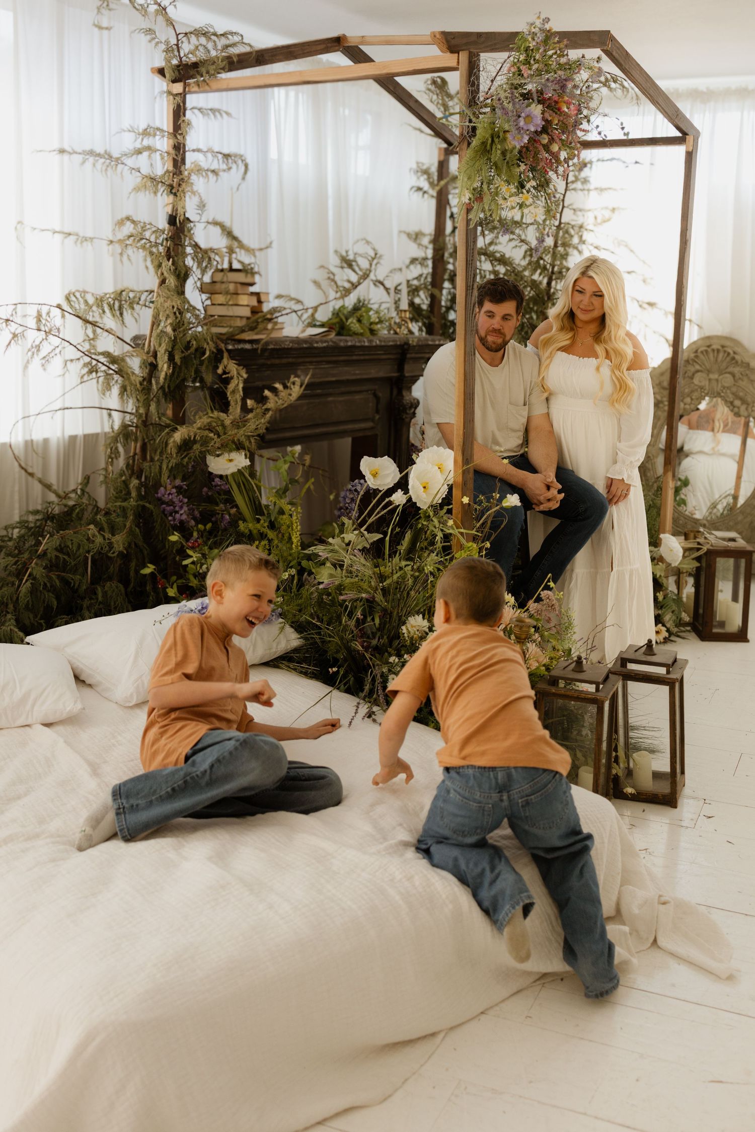 Pregnant woman in white dress with husband and two young boys playing on a canopy bed surrounded by lush greenery.