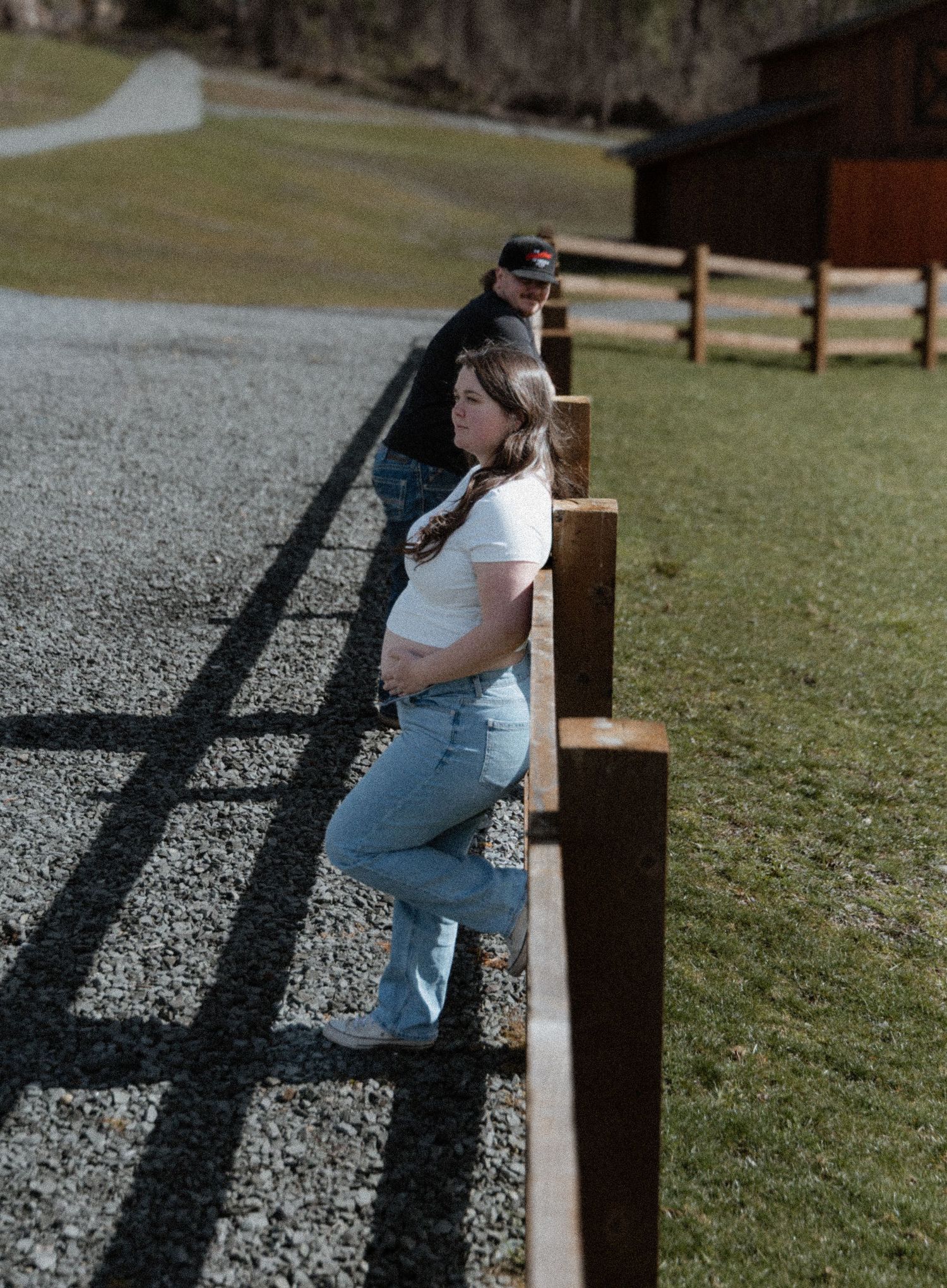 Person in blue jeans and white top leans against wooden fence post casting dramatic shadows on gravel and grass.