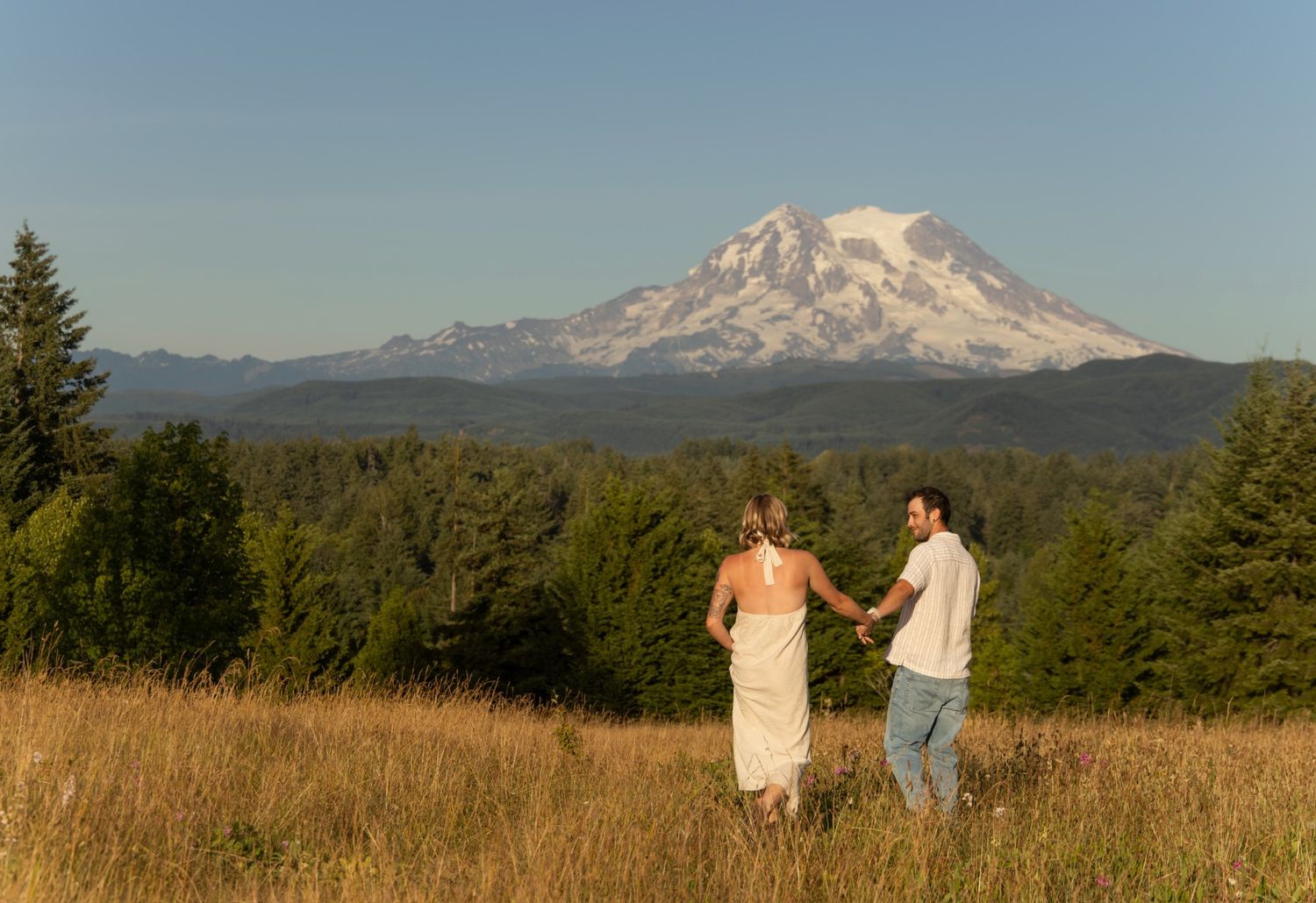 A panoramic view of Mount Rainier with two people walking through a golden meadow on a sunny day.