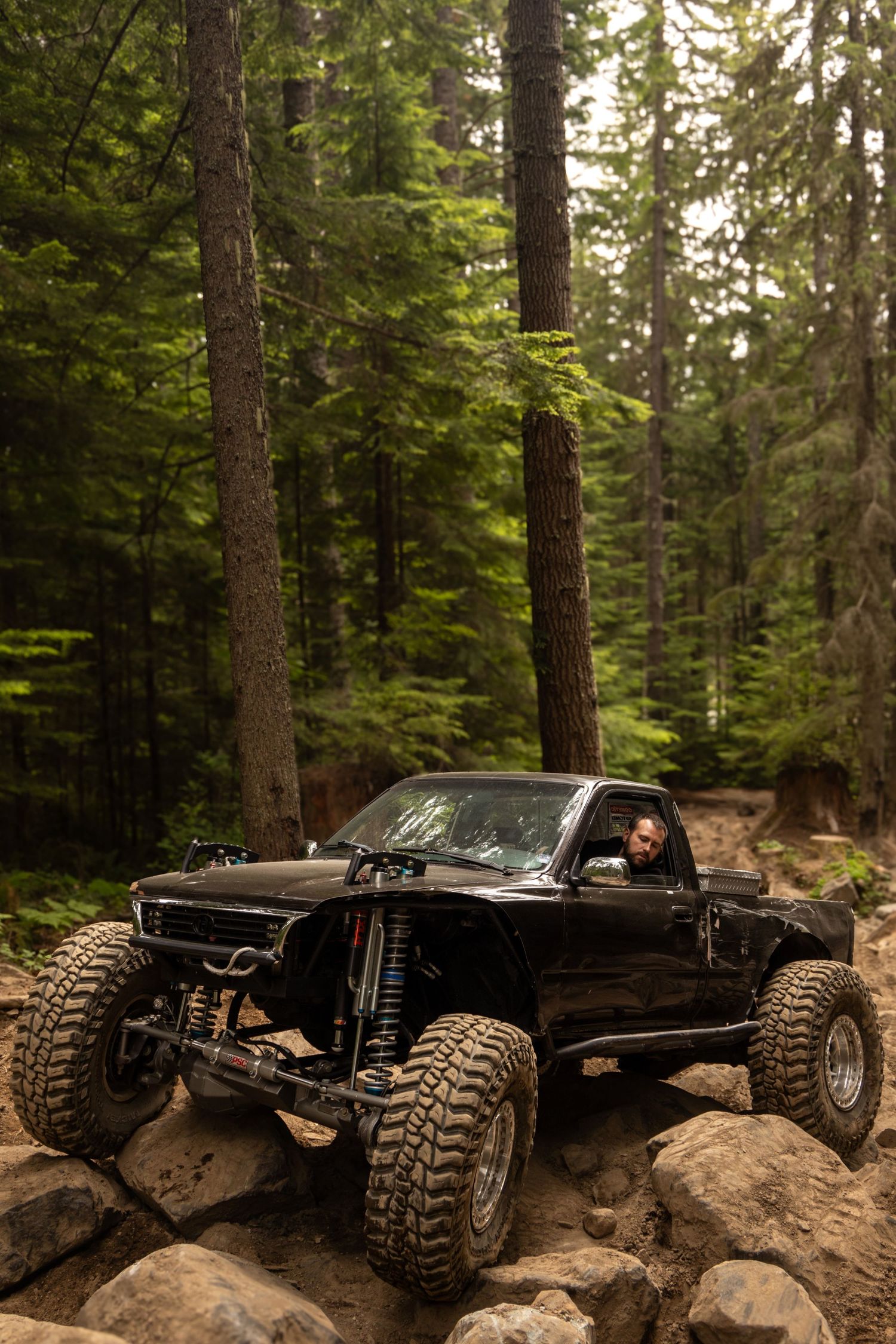 Black off-road vehicle navigating rocky terrain through dense forest on challenging mountain trail.