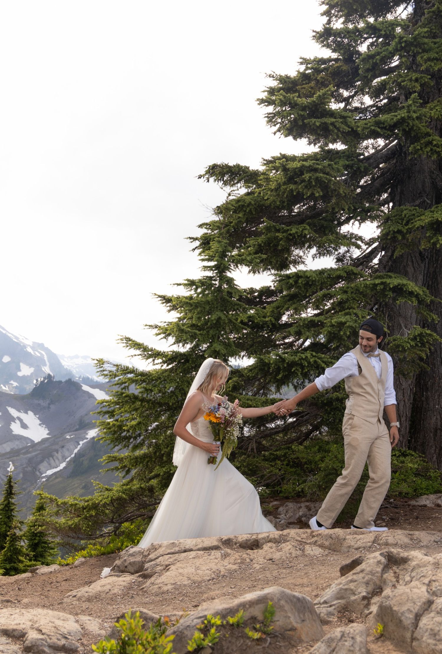 A couple poses for romantic wedding photos on a mountain ridge with evergreen trees and snow-capped peaks in the background.
