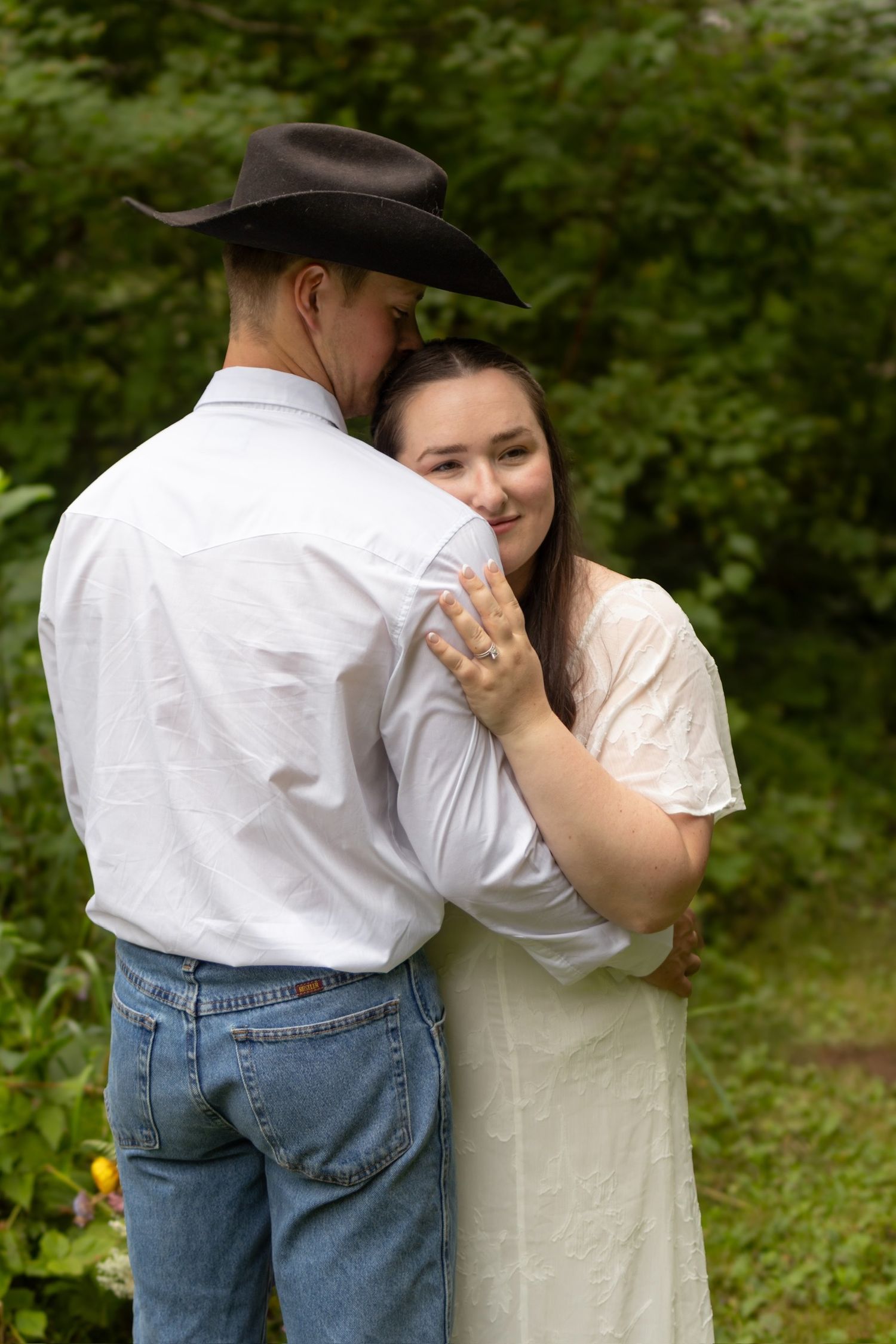 A couple in western attire share an intimate moment outdoors against a green natural backdrop.