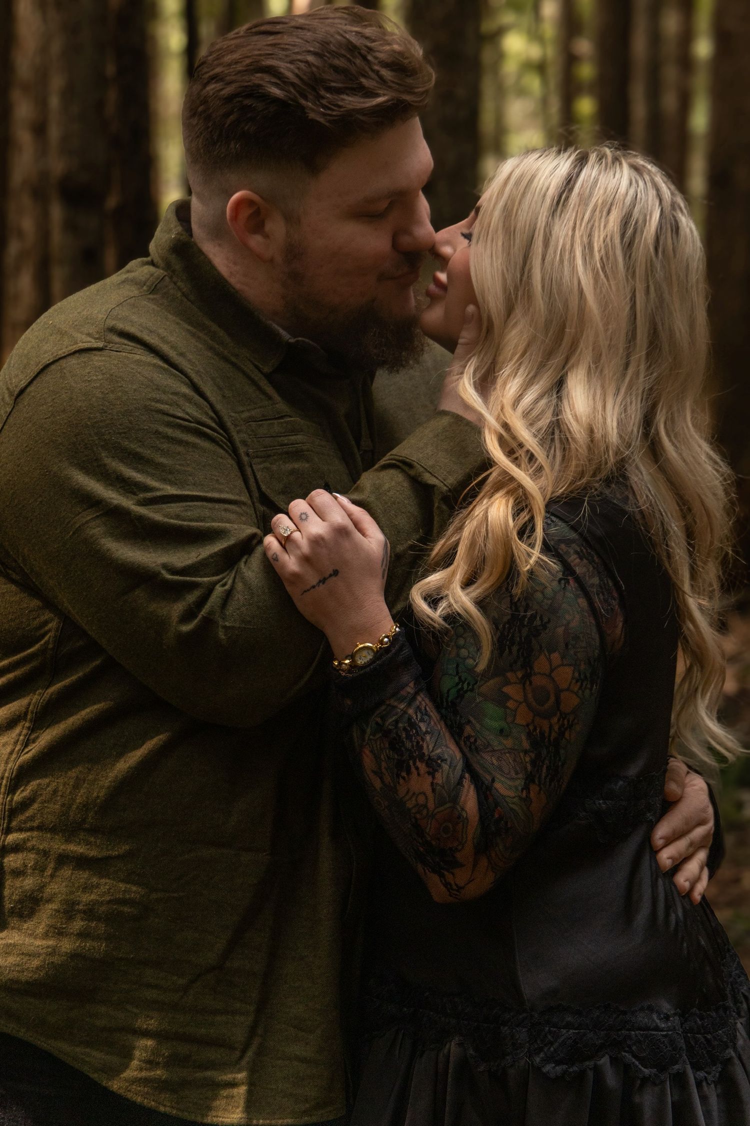 A romantic couple embraces in a forest setting with warm lighting and dark clothing.
