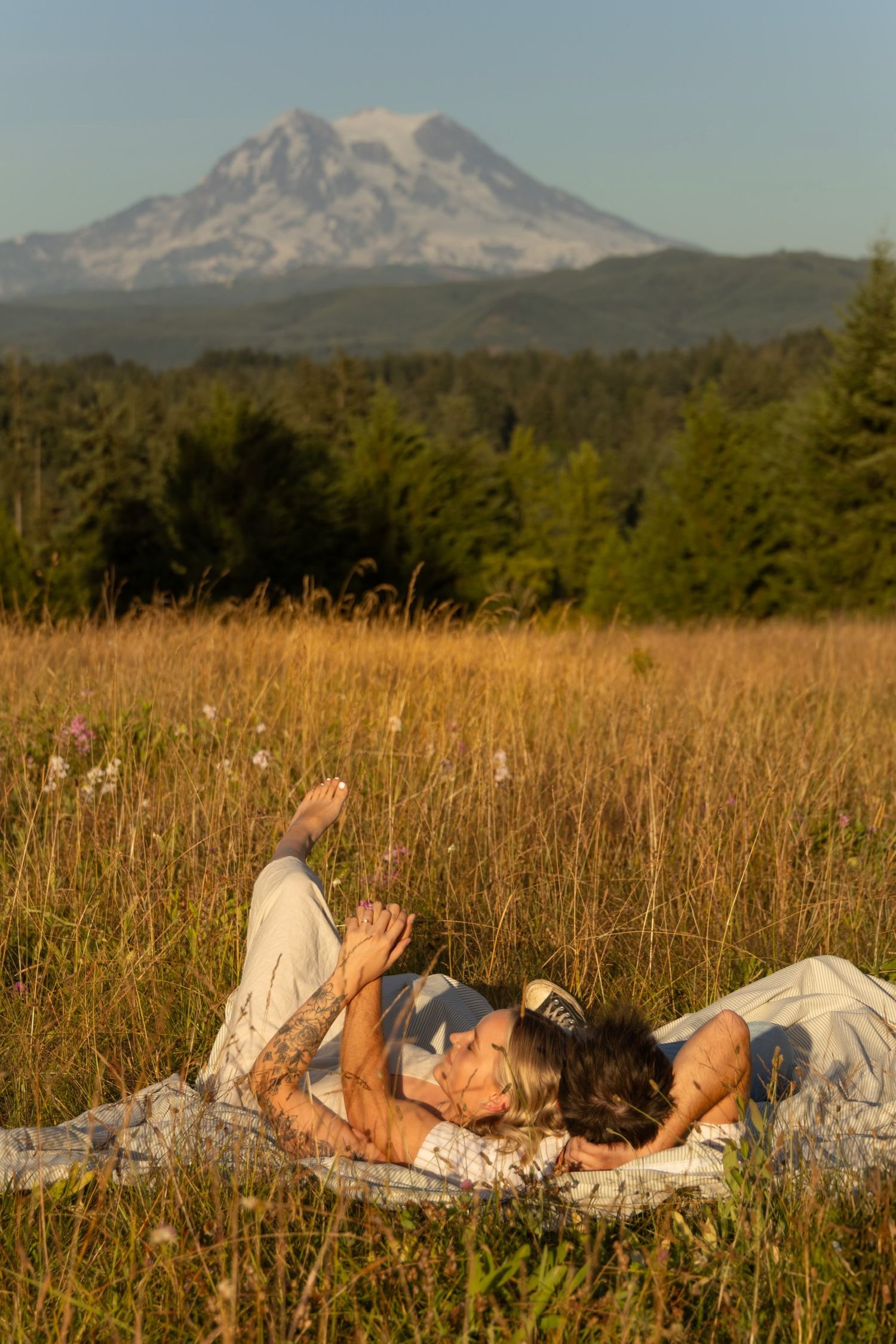 A couple lying on a blanket in a meadow with Mount Hood visible in the background during sunset.