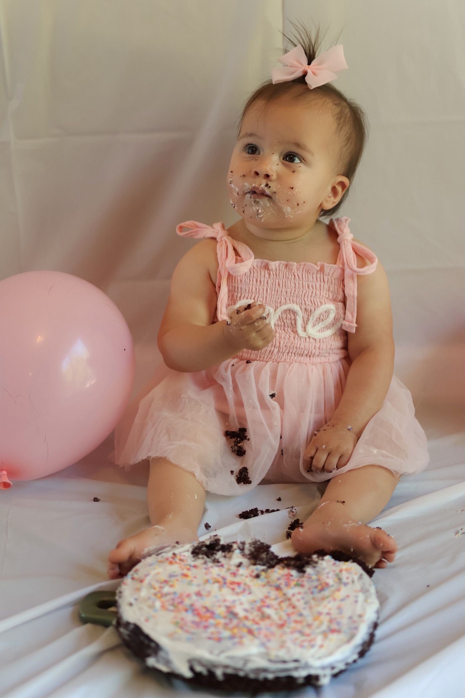 Baby in pink dress smashes birthday cake with white frosting and sprinkles next to pink balloon on white backdrop.