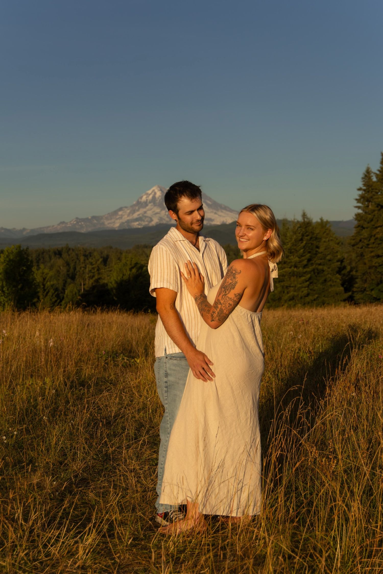 A couple embraces in a golden field at sunset with mountain peaks visible in the background.